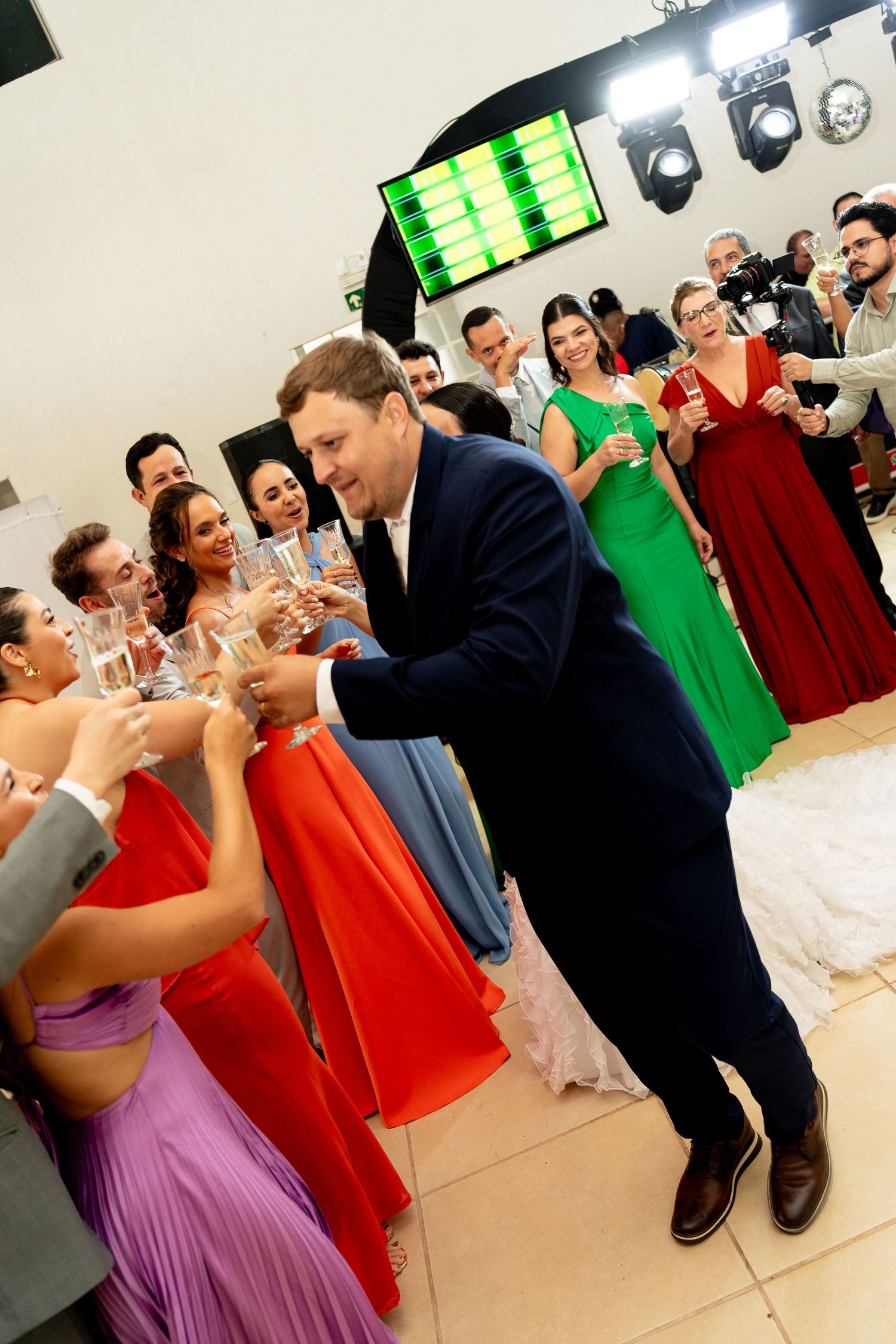 Groom in navy suit toasting with wedding guests in colorful gowns at a festive reception celebration.
