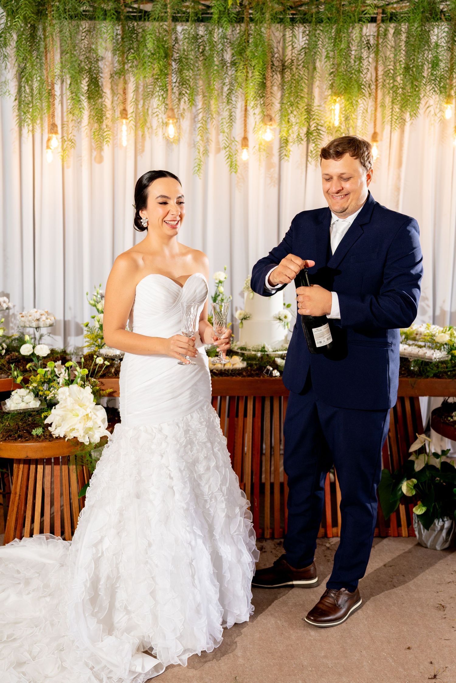 Bride in white gown and groom in navy suit open champagne bottle at elegant wedding reception with floral decor.