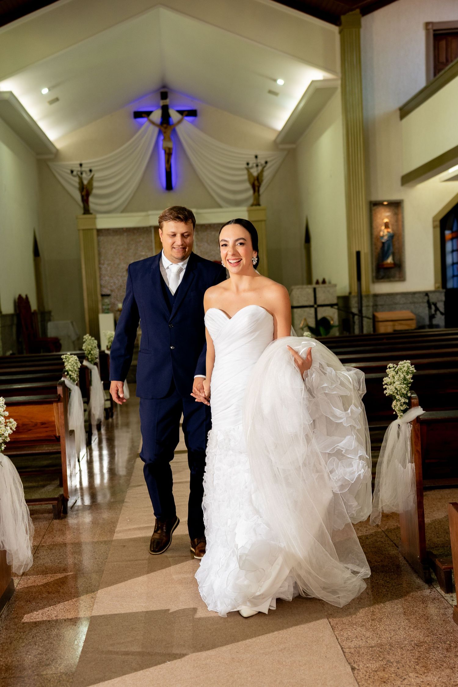 Smiling bride in white gown and groom in navy suit walk down church aisle after wedding ceremony.