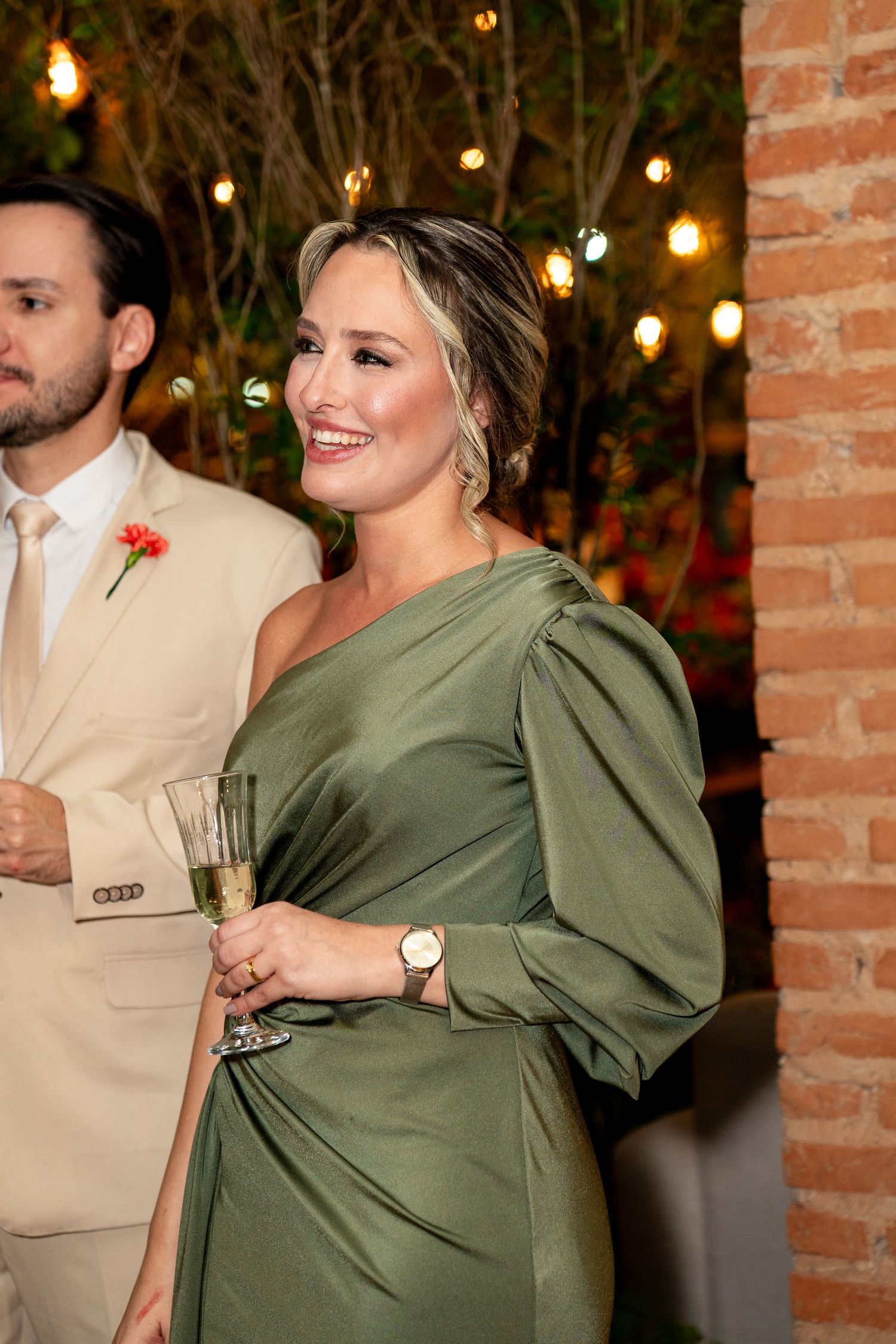 A smiling woman in an elegant olive green one-shoulder gown holds a champagne glass at a wedding reception.