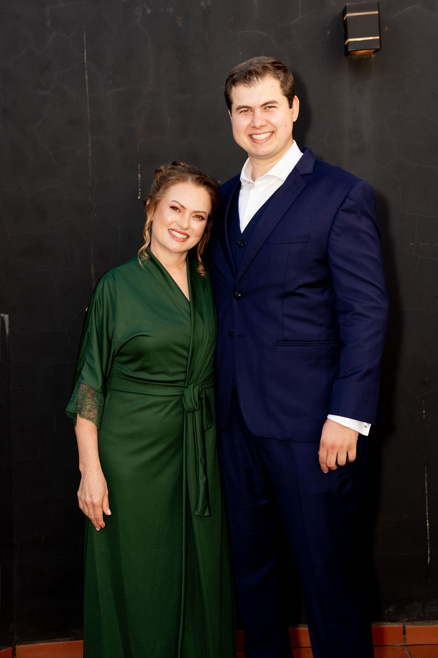 A smiling couple poses together, woman in green dress and man in navy blue suit, against a dark wall.
