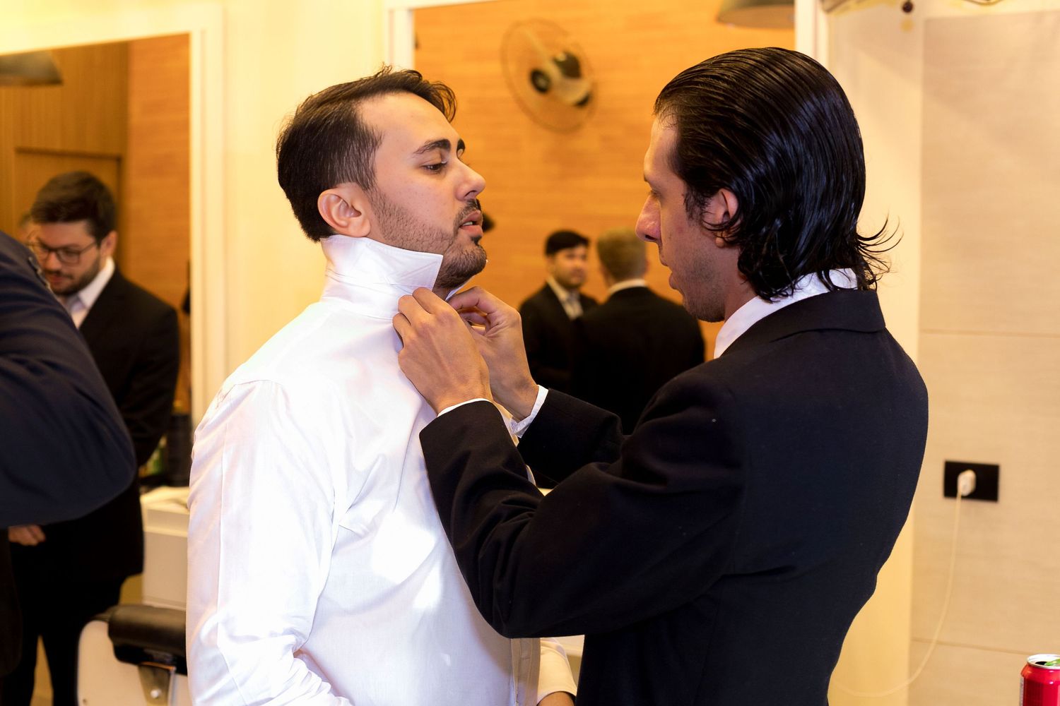 Two men in formal attire, one adjusting the other's collar in an elegant indoor setting.