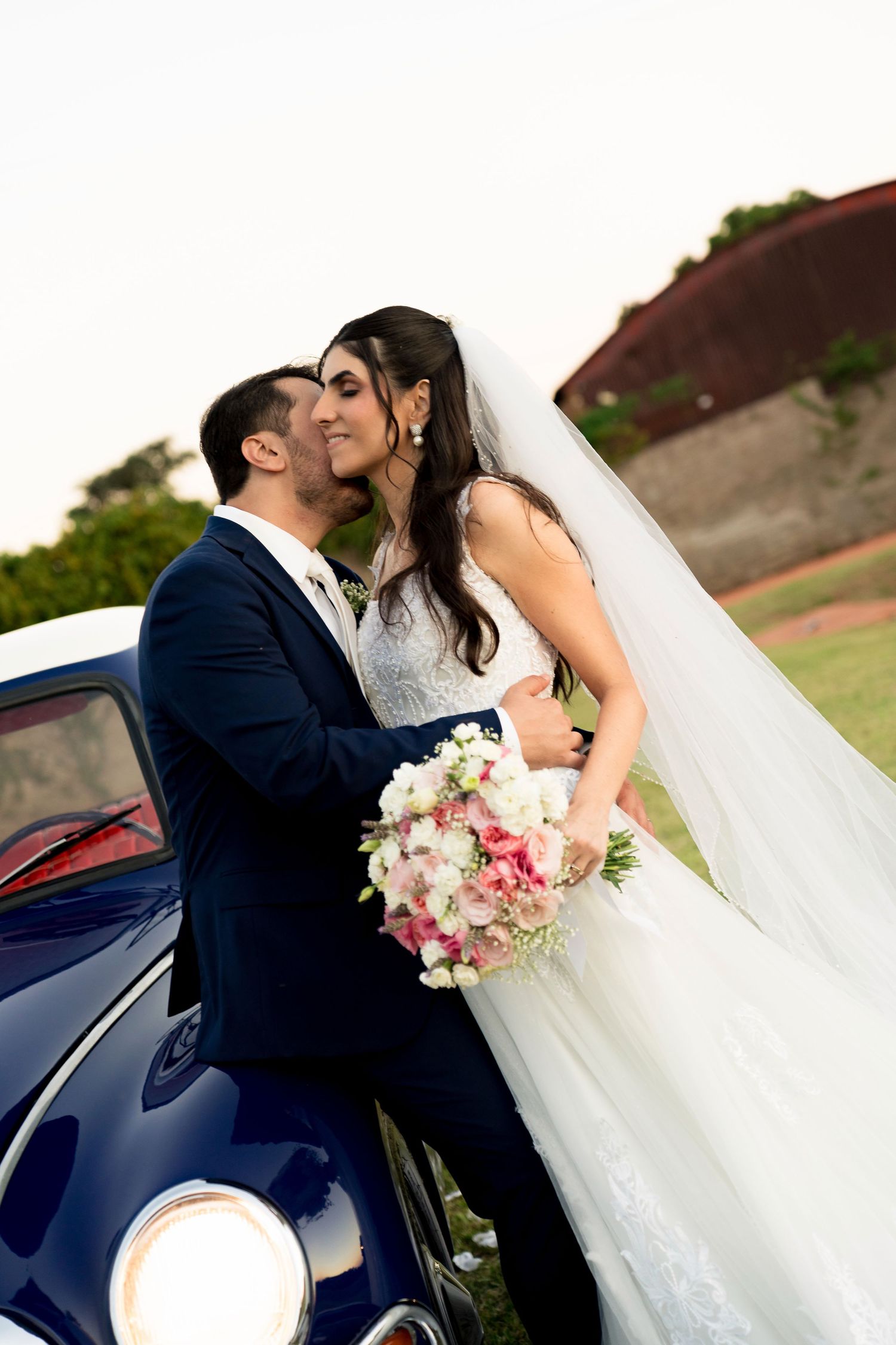 Bride and groom share a romantic kiss leaning against a vintage blue car, bride holding a pink and white bouquet.