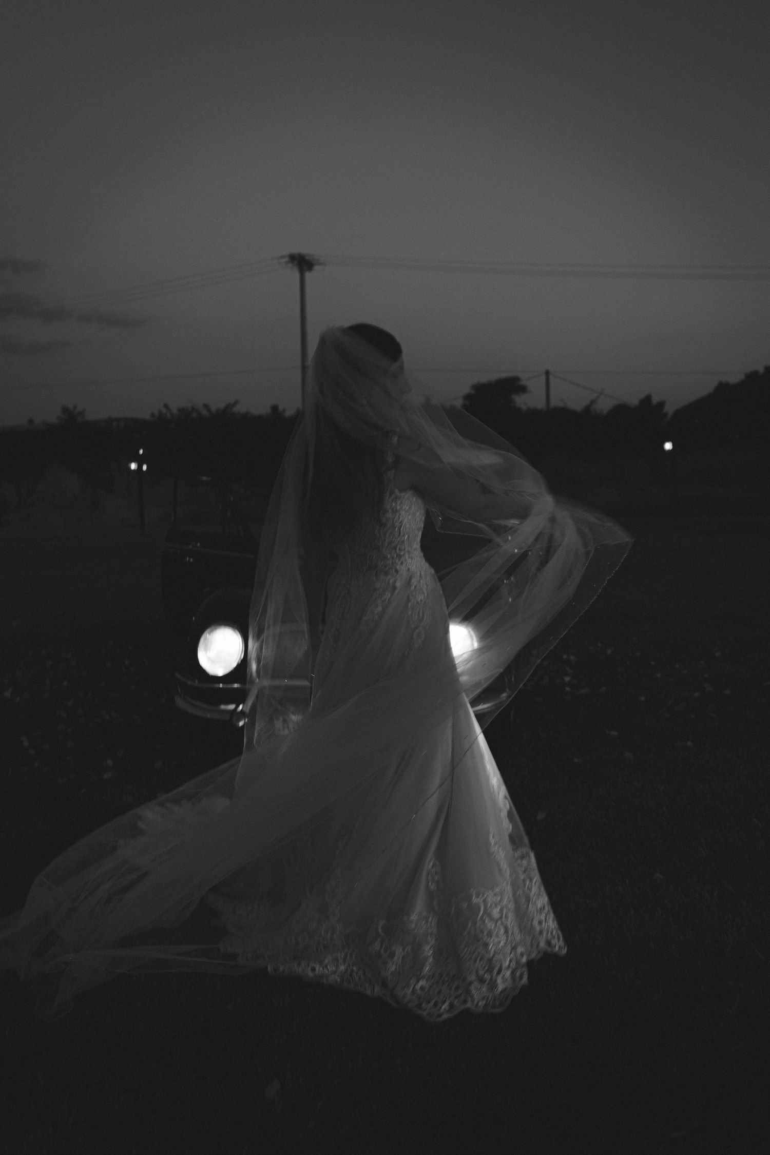 Black and white photo of a bride in lace gown and flowing veil standing before vintage car headlights at dusk.