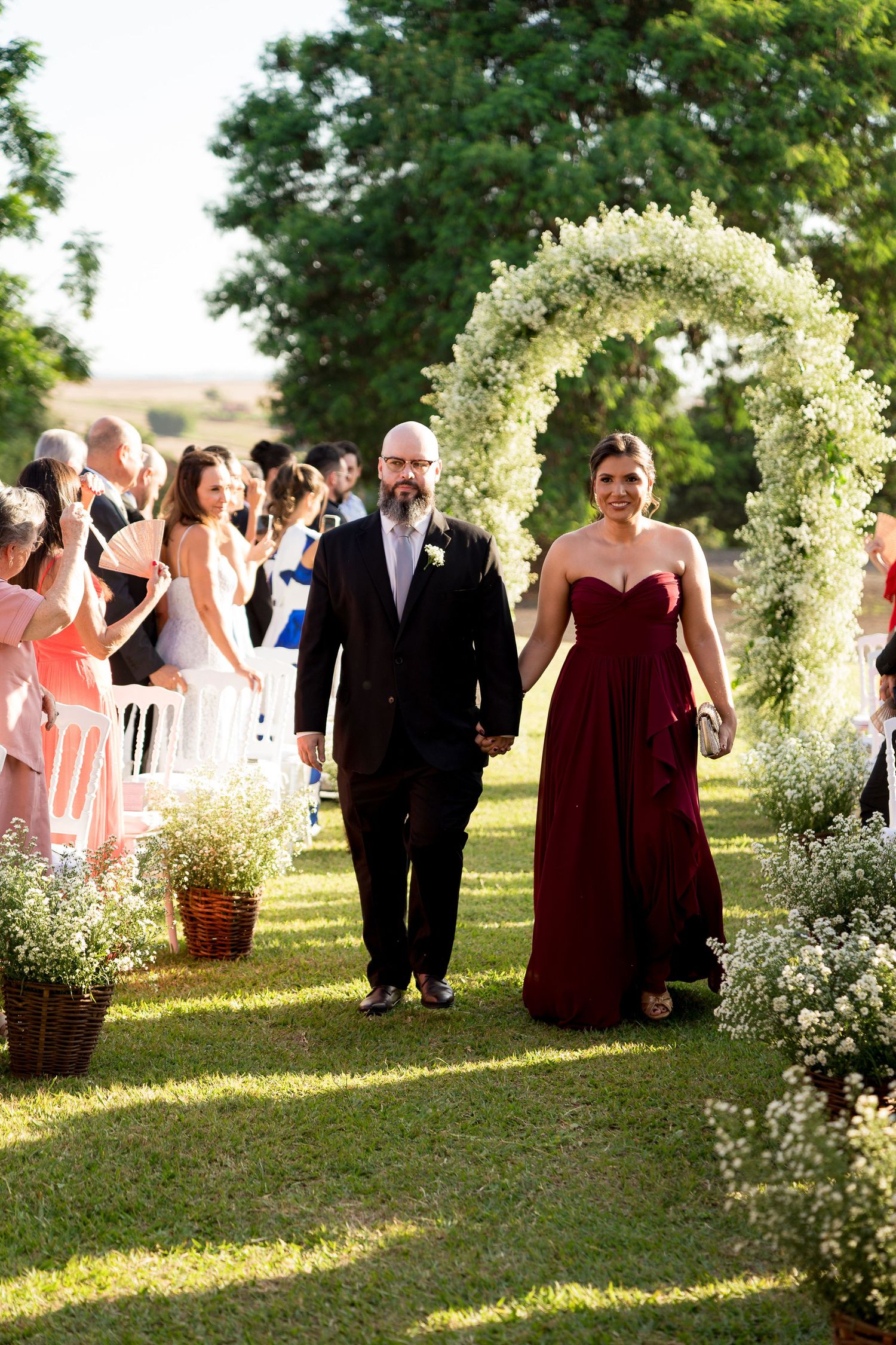 Couple walks down outdoor wedding aisle under floral arch, woman in burgundy gown, man in dark suit, guests applauding.
