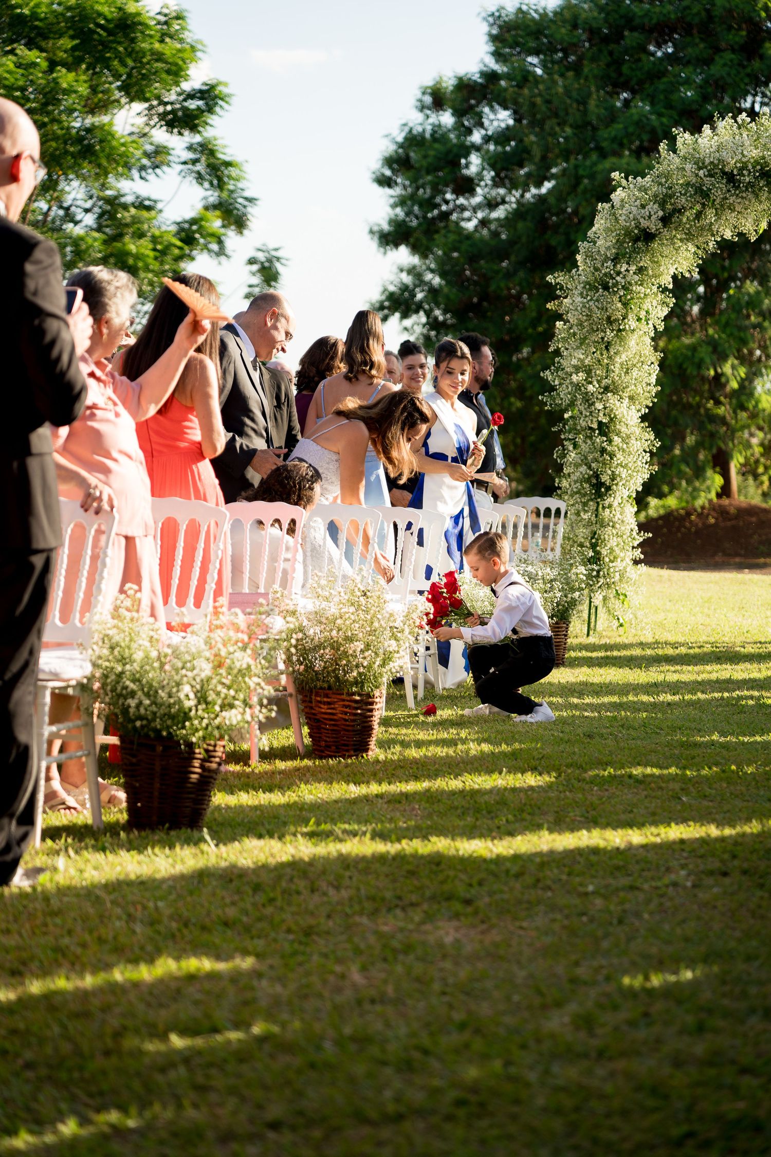 Outdoor wedding ceremony aisle with guests seated on white chairs, floral arch, and a child crouching near red roses.