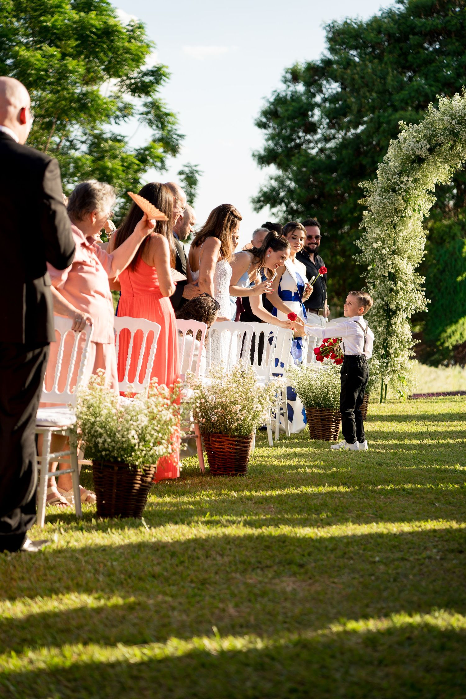 Outdoor wedding ceremony on green lawn with guests seated in white chairs, floral arch, and a young ring bearer walking the aisle.