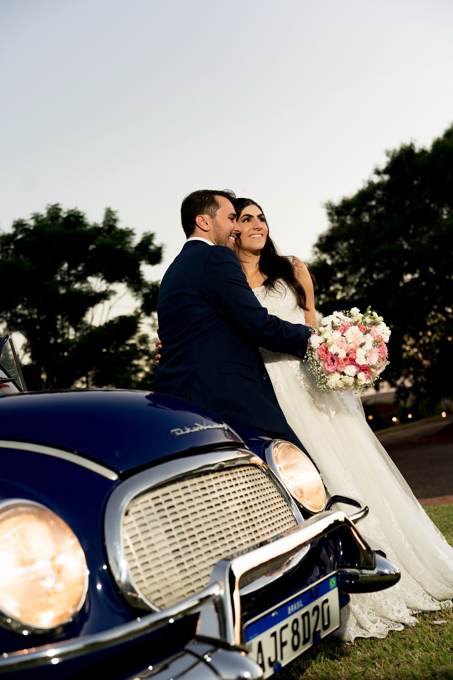 Bride and groom pose romantically on a vintage classic car, bride holding pink bouquet, at dusk outdoor wedding.