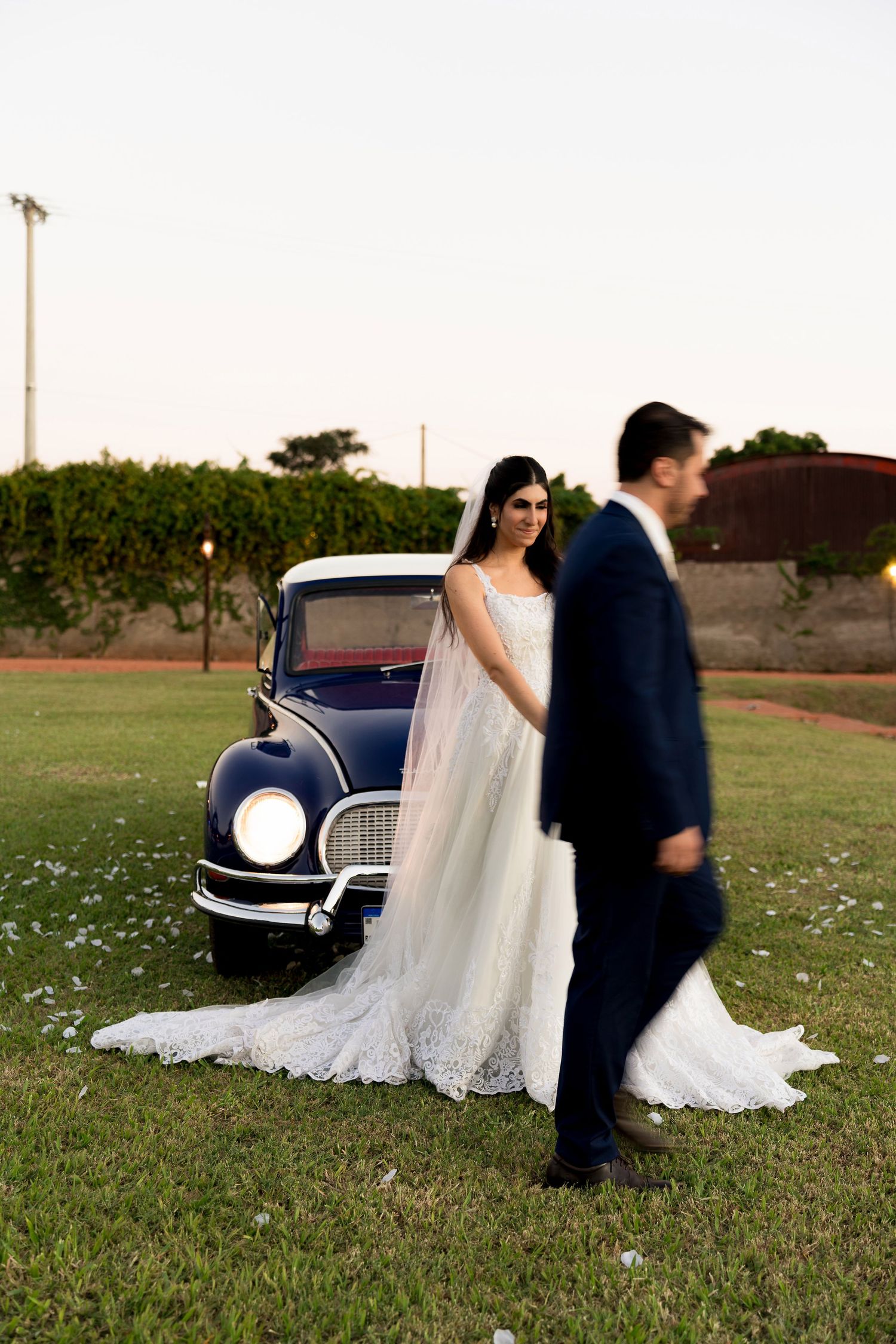 Bride in lace gown stands by vintage navy car as groom in suit walks past on green lawn at dusk.
