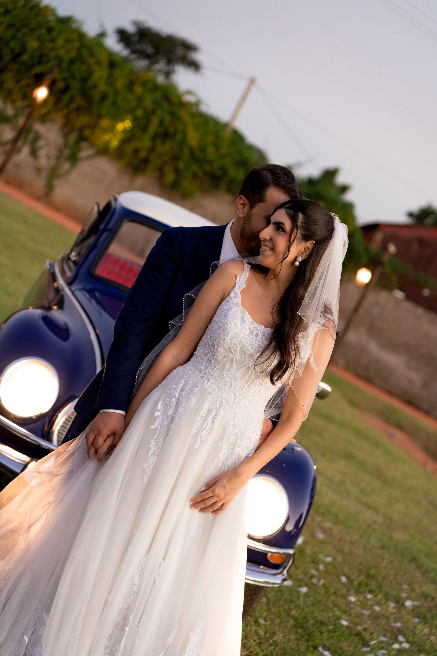 A bride and groom share a romantic moment leaning against a vintage blue car at dusk during their outdoor wedding.