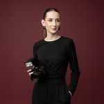 A young woman in a black long-sleeve dress holds a camera, posing confidently against a dark red background.