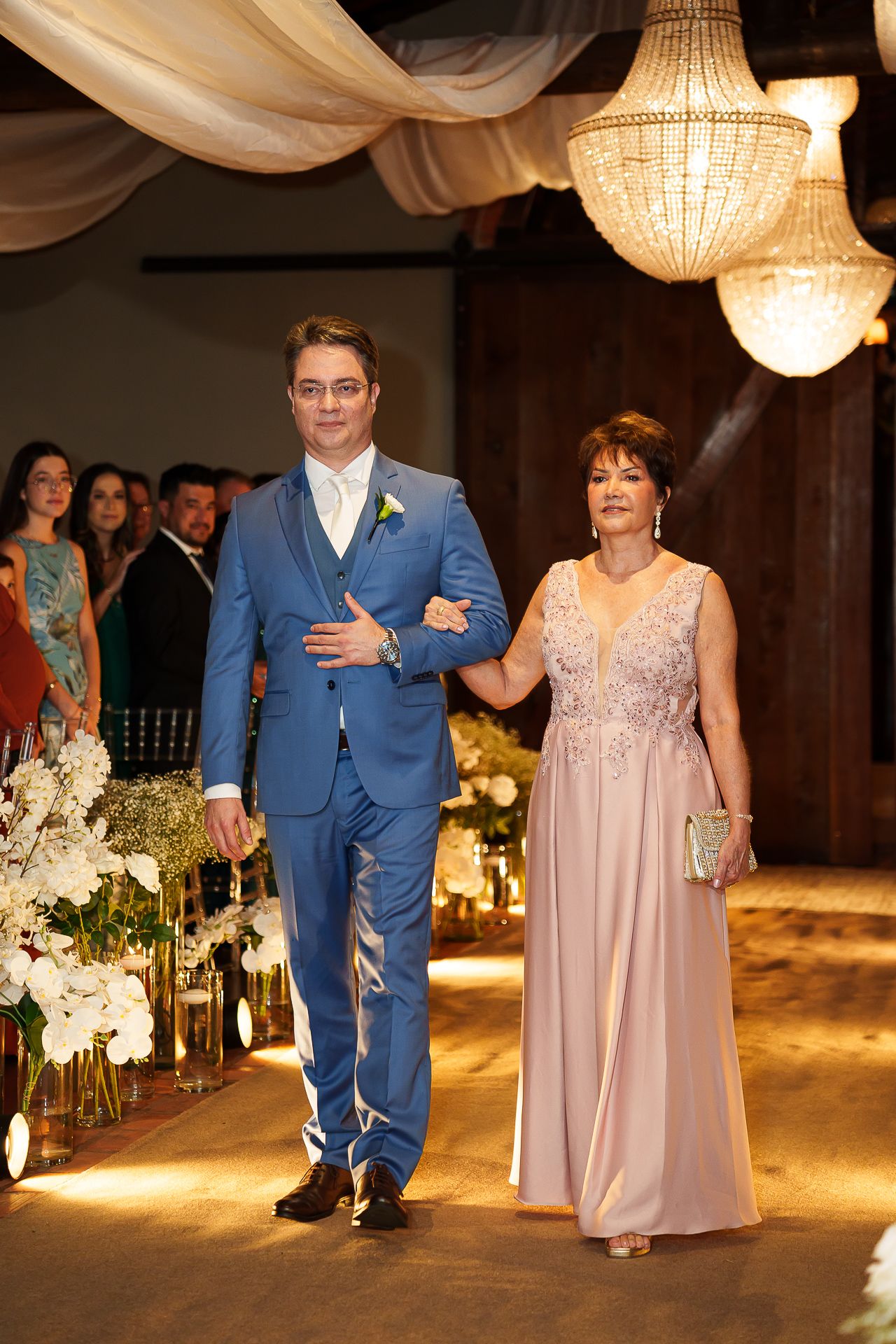 Groom in blue suit escorts elegantly dressed woman in pink gown down wedding aisle under crystal chandeliers.