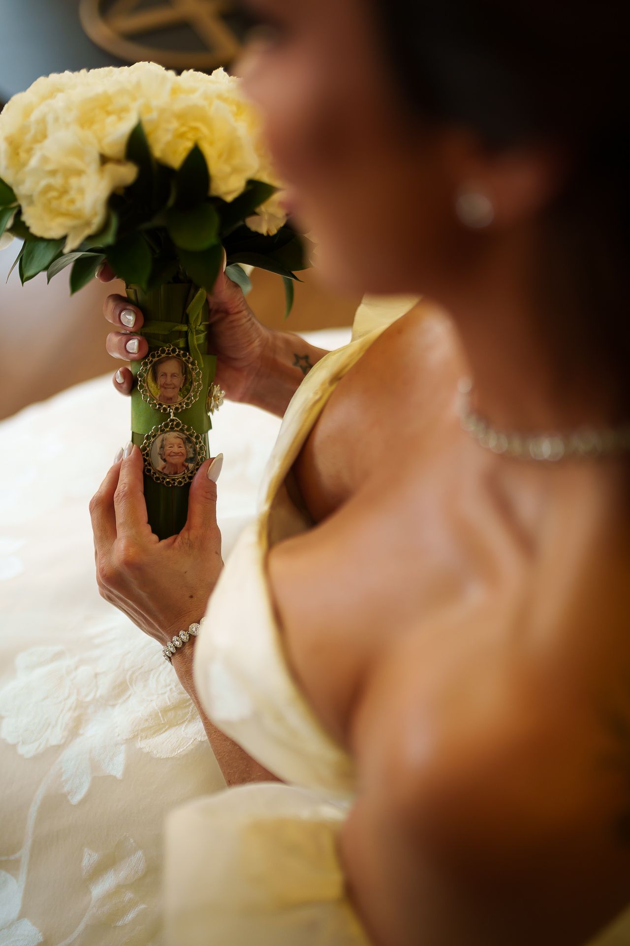 Bride in white gown holding yellow rose bouquet with memorial photo charms on stems at wedding ceremony.