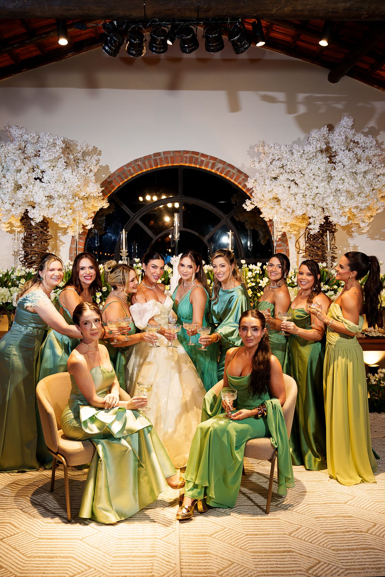 Bride in white gown poses with bridesmaids in green satin dresses, holding champagne glasses at elegant floral wedding reception.