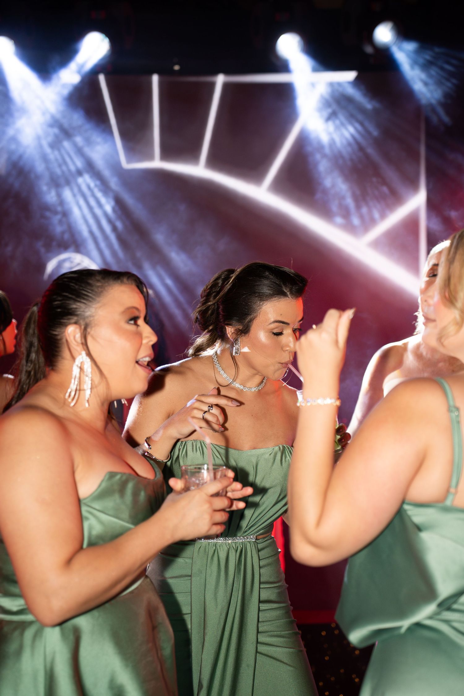Three women in green satin dresses dancing and laughing together under stage lights at a formal event.