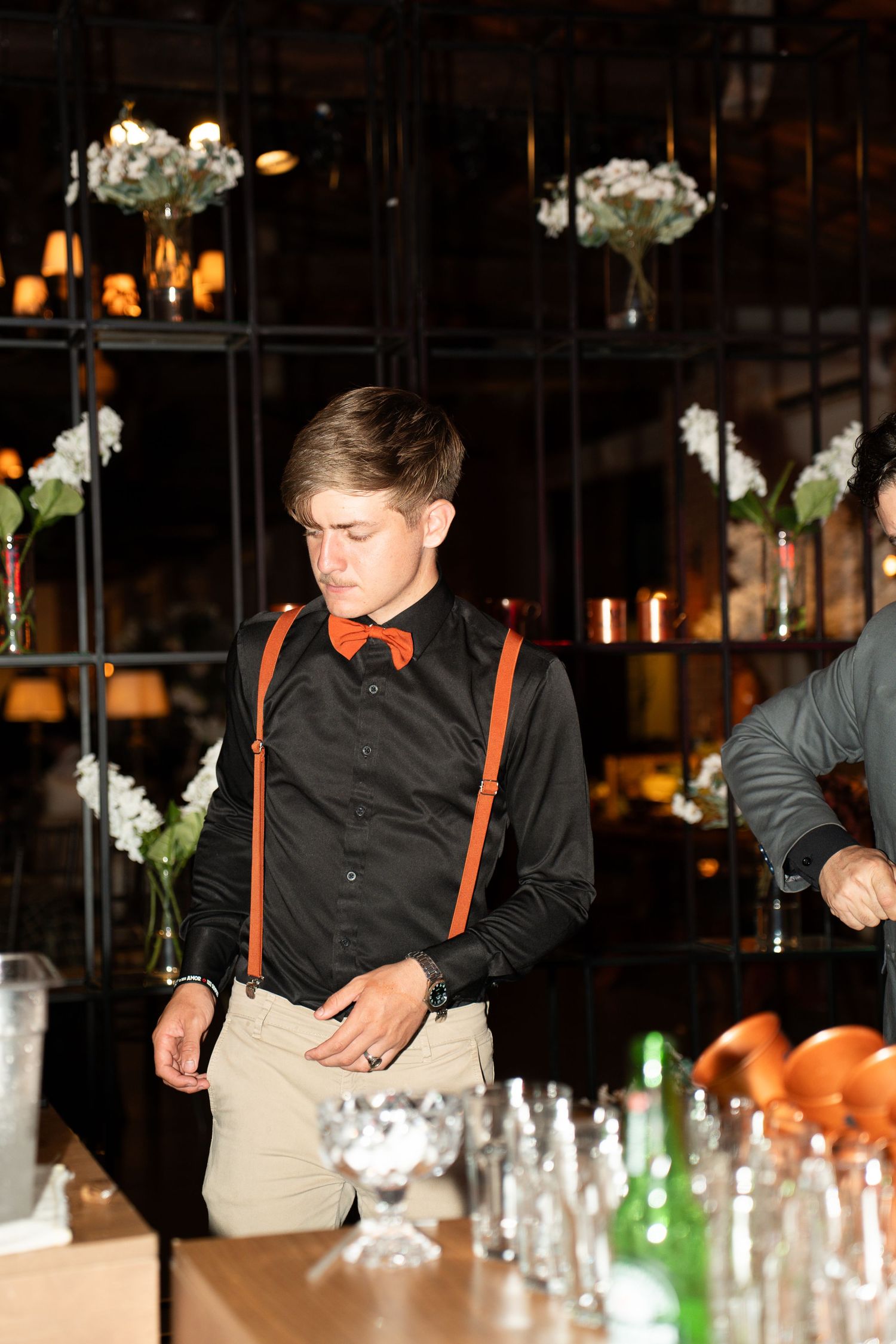 Young bartender in black shirt, orange bow tie and suspenders standing behind a bar with glasses and bottles.