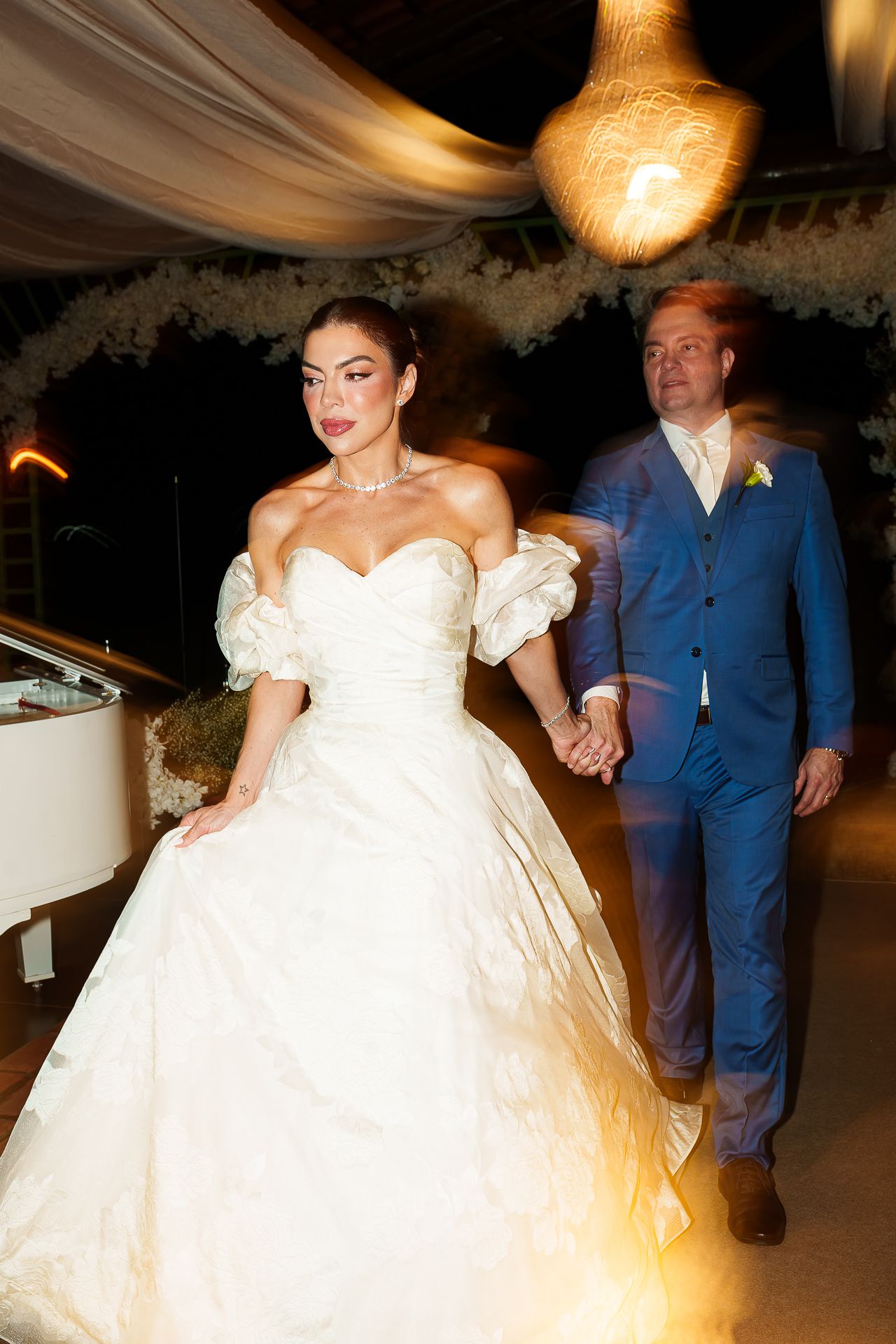 Bride in white off-shoulder ball gown holds hands with groom in blue suit during wedding reception dance.