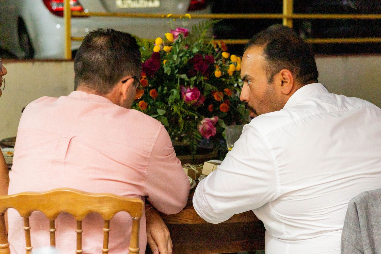 Two men in pink and white shirts sitting together at a table with colorful flower arrangement at event.