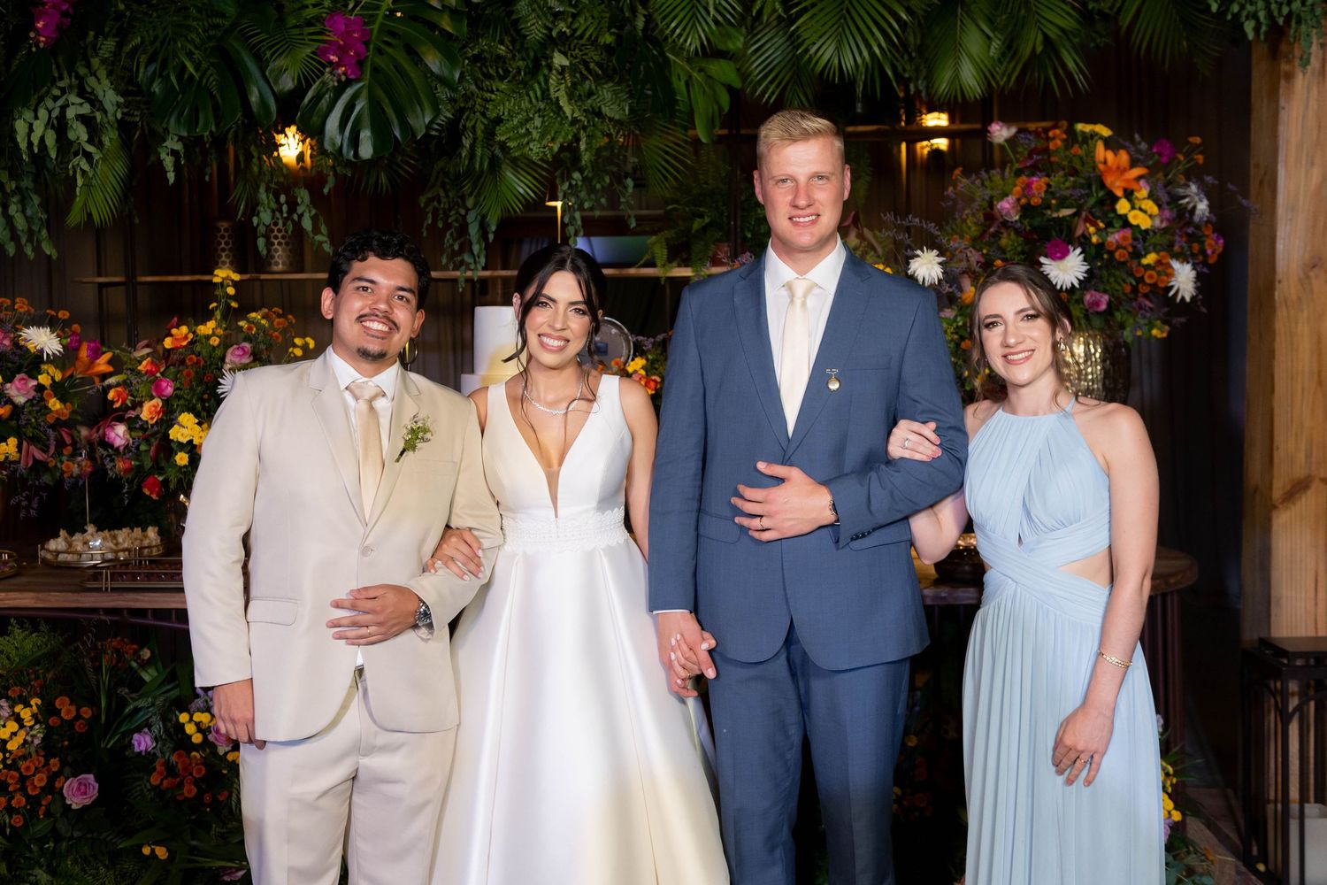 Wedding party of four posing indoors with tropical floral backdrop, bride in white gown, groom in blue suit.