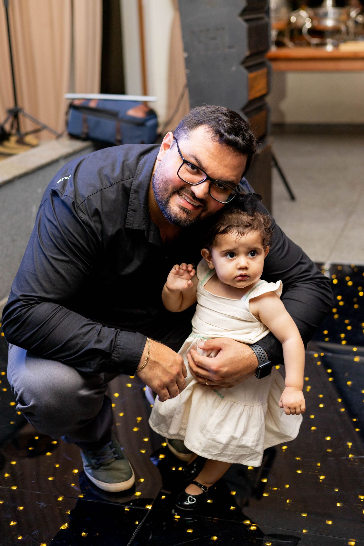 Man with glasses crouching beside a toddler girl in white dress on a dark floor with gold lights.
