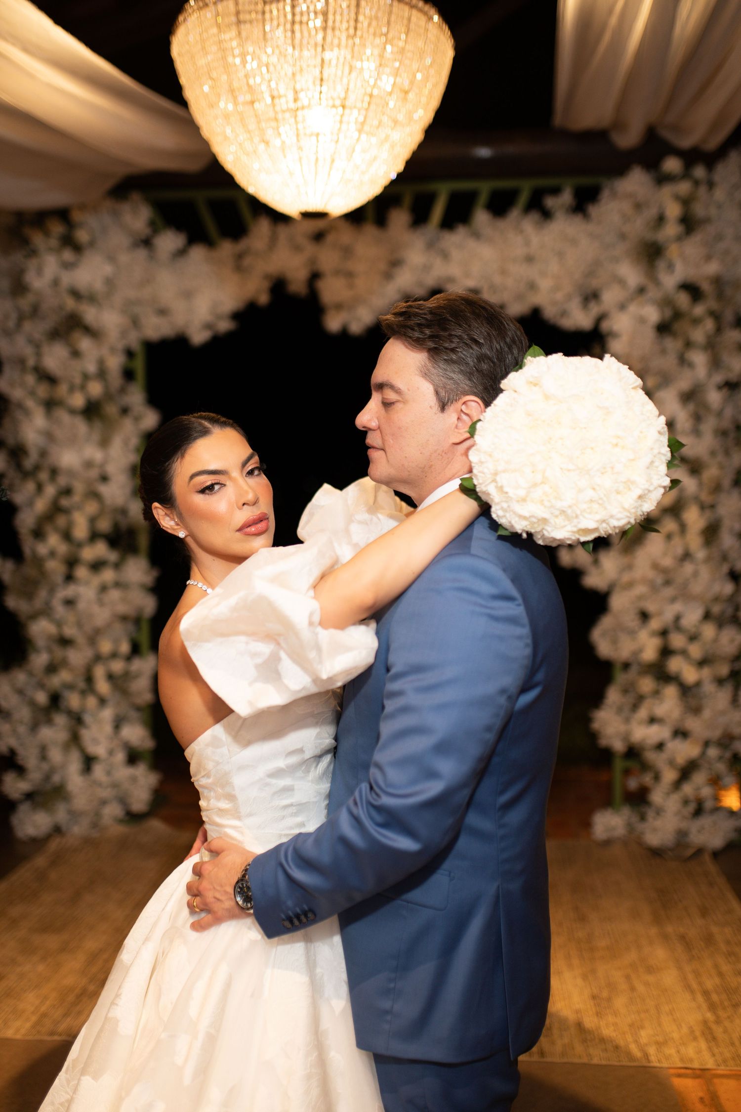 Bride and groom share a romantic dance at their elegant wedding reception with white floral backdrop and chandelier lighting.