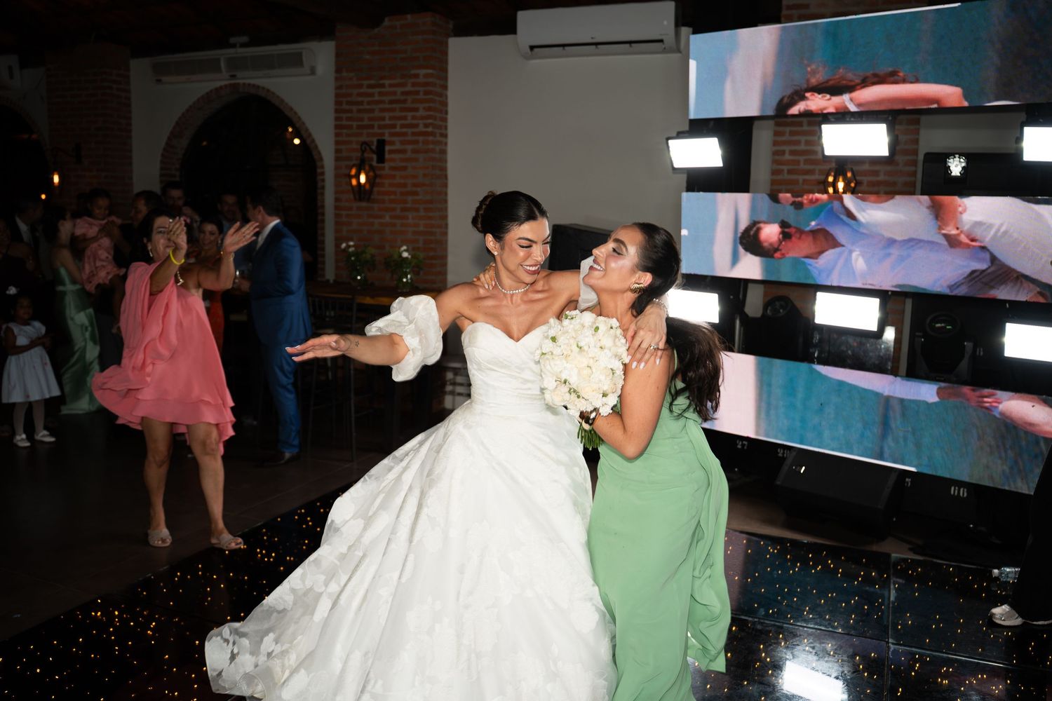 Bride in white gown hugs bridesmaid in green dress on dance floor at elegant wedding reception.