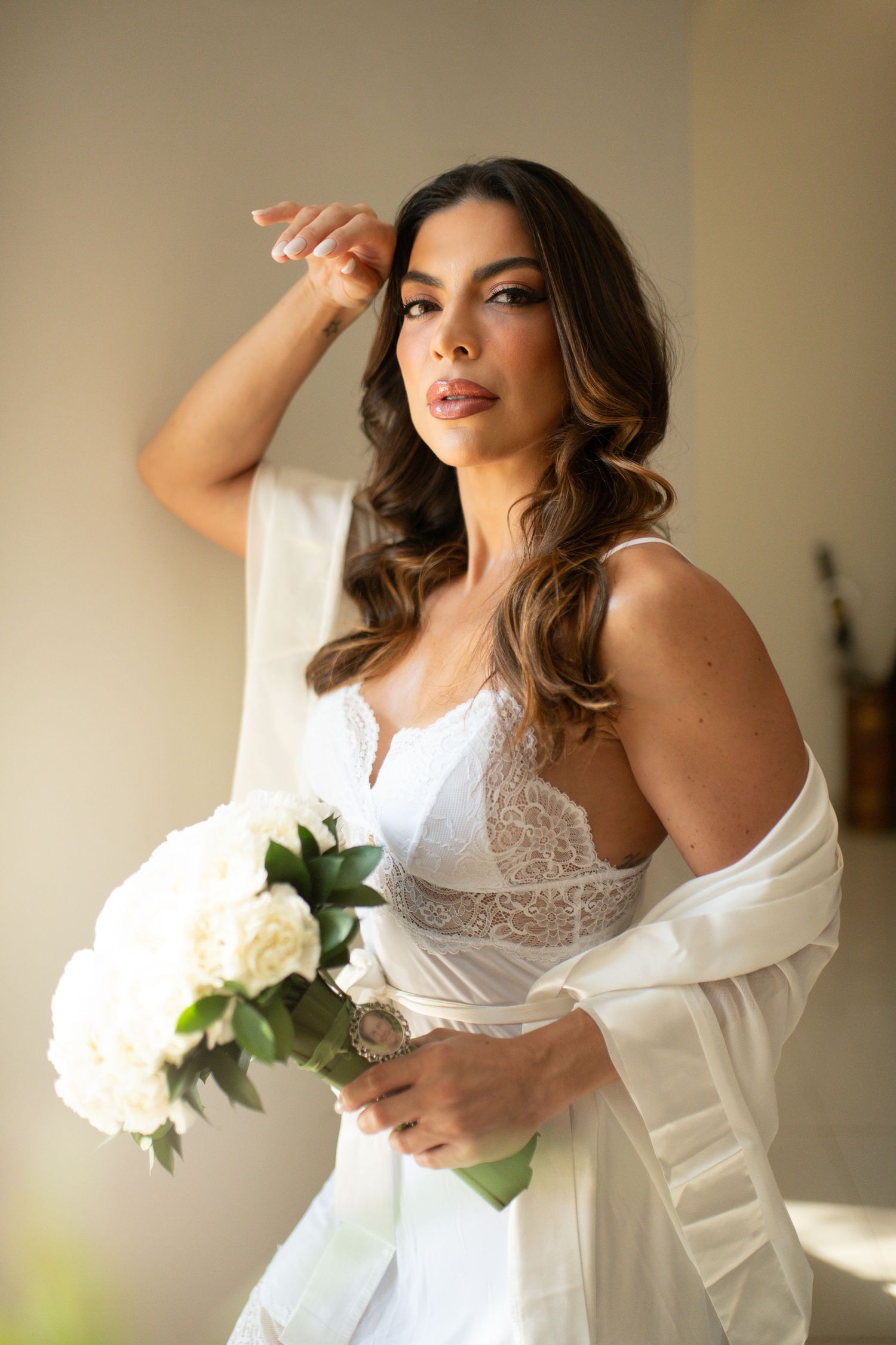 Bride in white lace bodice and sheer robe holding white peony bouquet, posing elegantly in soft natural light.