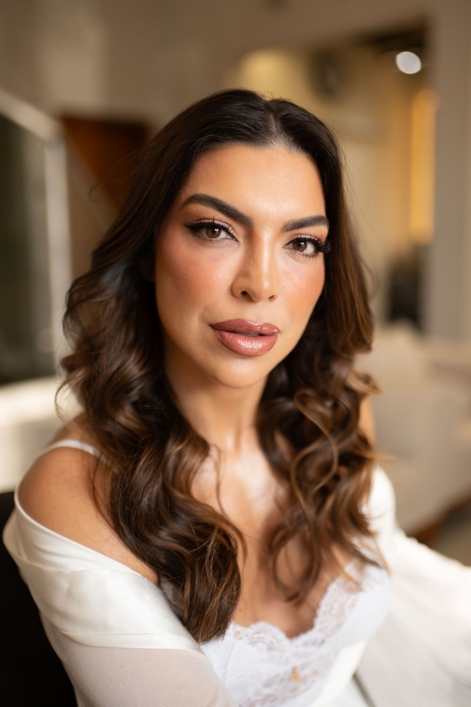 A woman with curly brunette hair and elegant makeup wearing a white off-shoulder lace top poses for a bridal portrait.