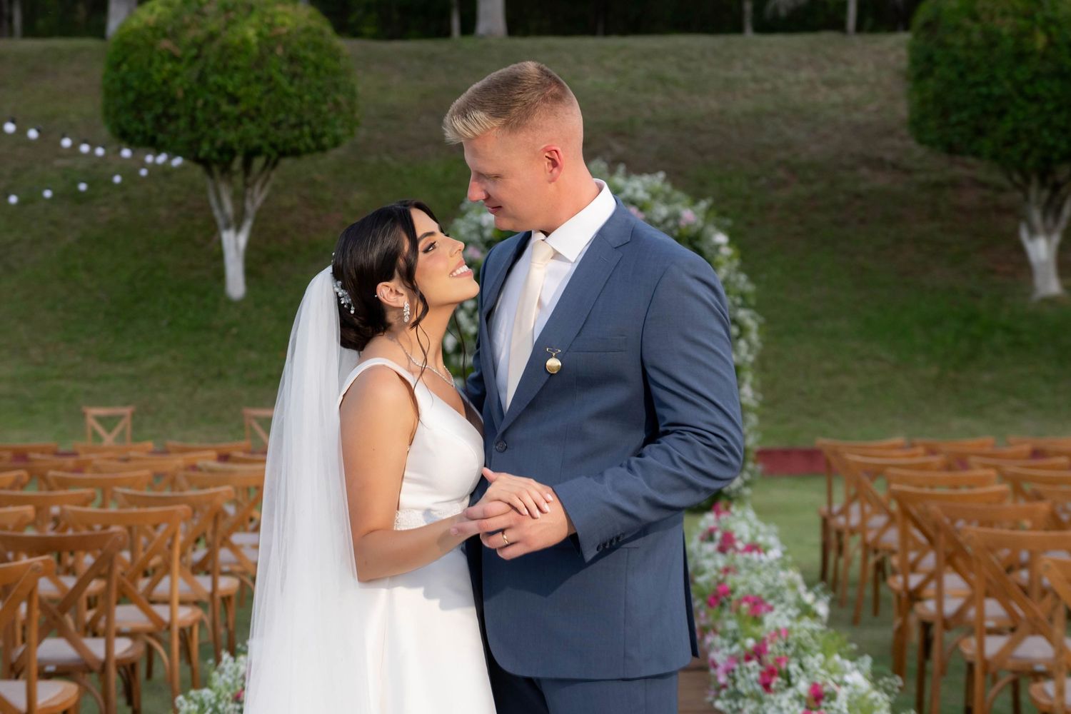Bride in white gown and groom in blue suit share a romantic moment at an outdoor wedding ceremony.