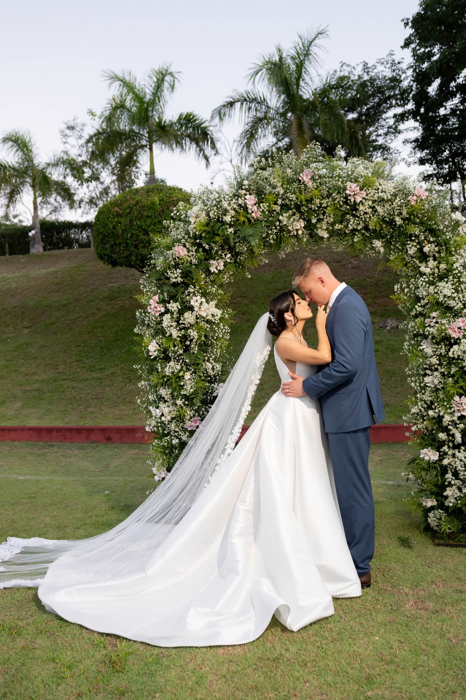 Newlyweds kiss beneath a lush floral arch with palm trees in the background at an elegant outdoor wedding ceremony.