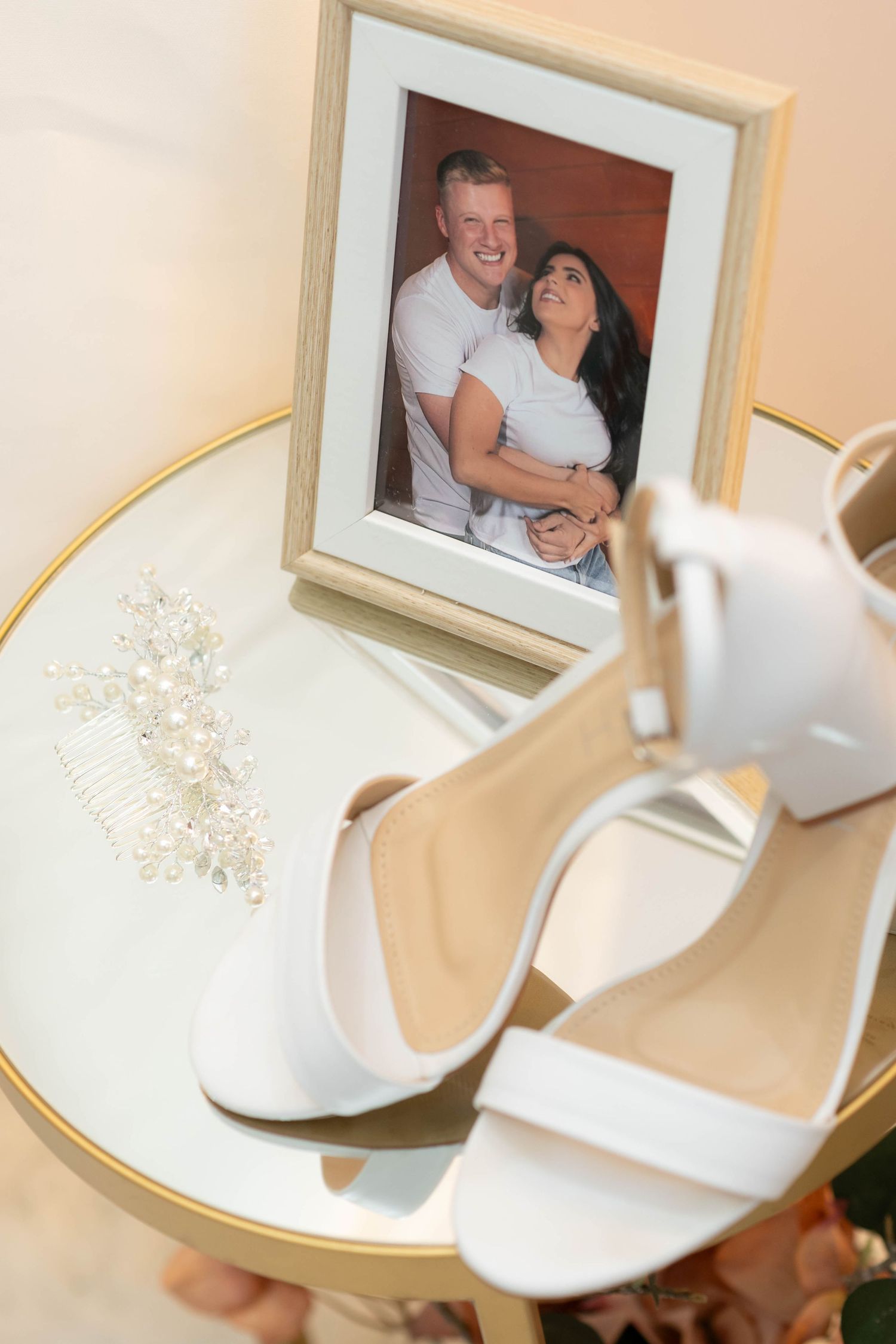 White bridal heels and pearl hair comb displayed on gold mirror table with framed couple photo.