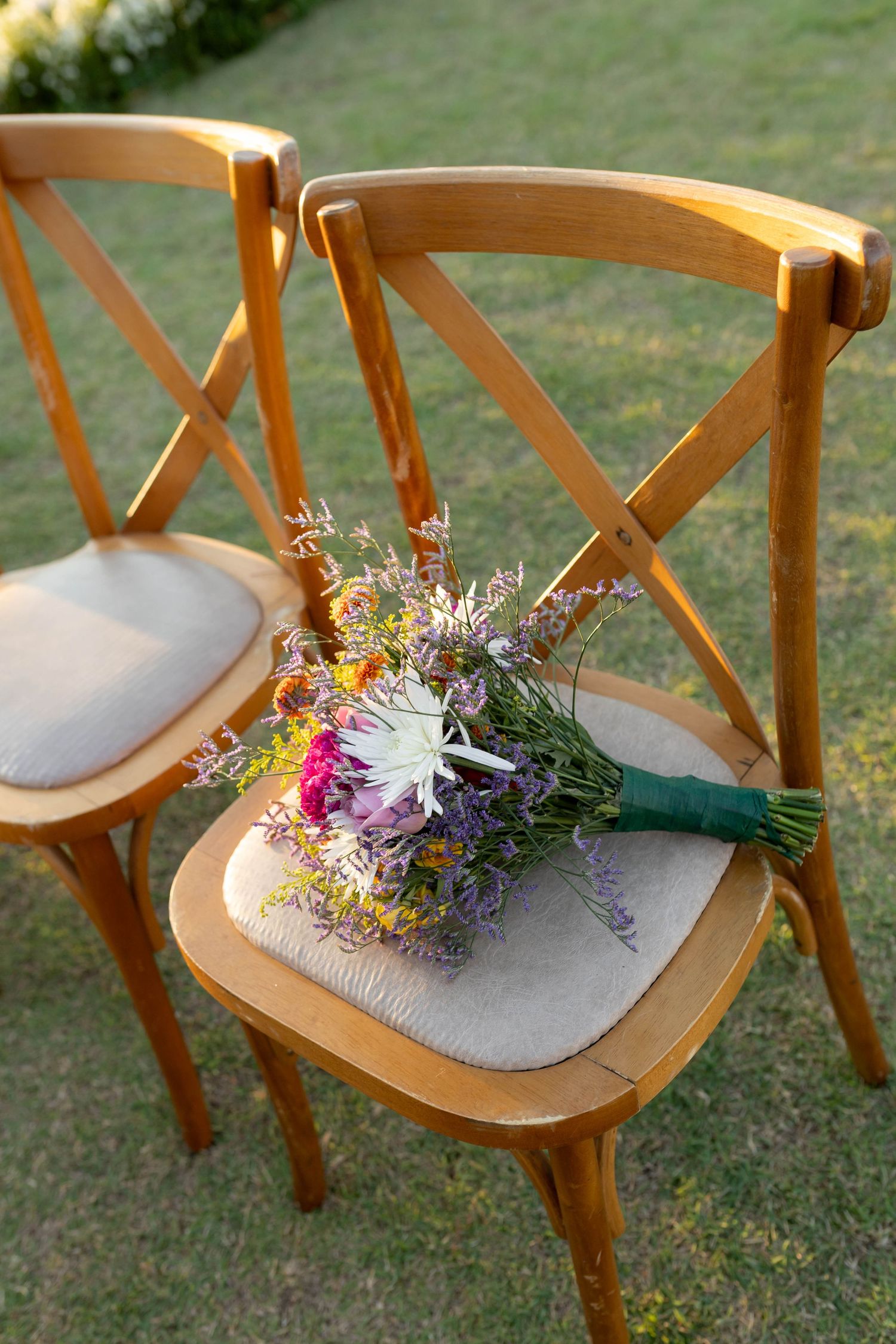 Colorful wildflower bridal bouquet wrapped in green ribbon resting on wooden cross-back chair on green grass.