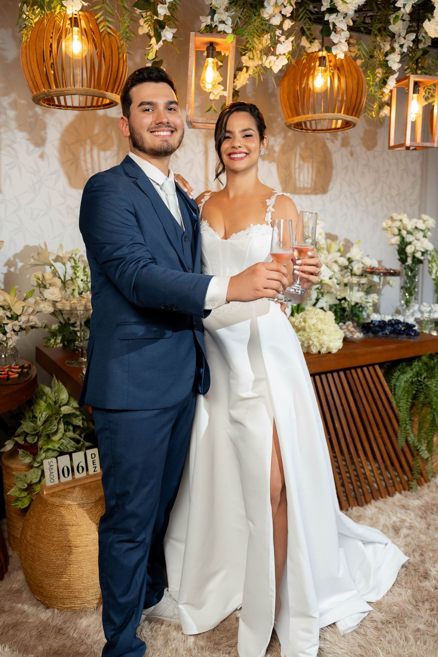 Smiling bride and groom in wedding attire toasting with champagne glasses amid floral decorations and warm pendant lights.