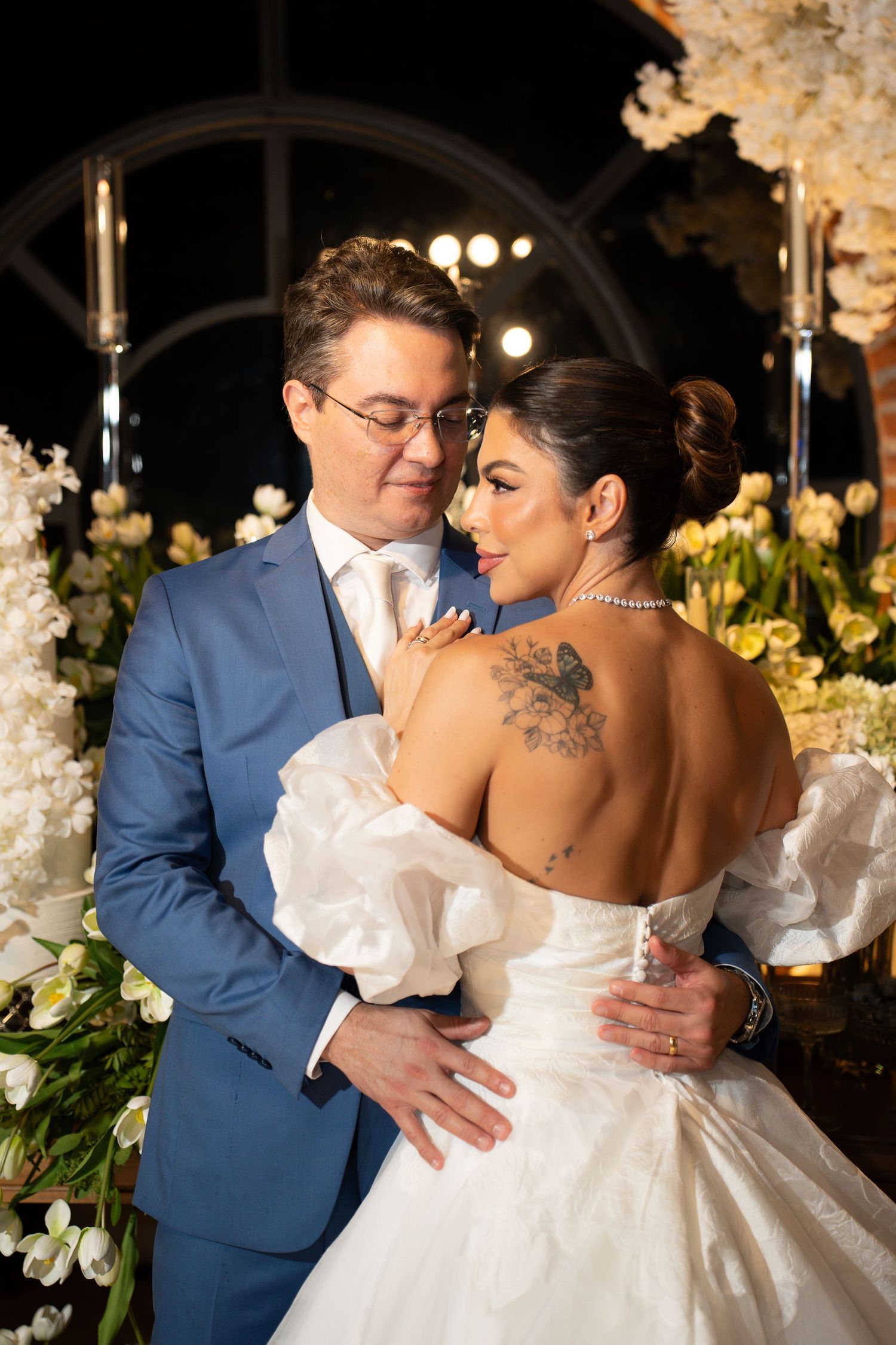 Bride and groom embrace at elegant wedding ceremony, bride in backless gown with floral tattoo, groom in blue suit.