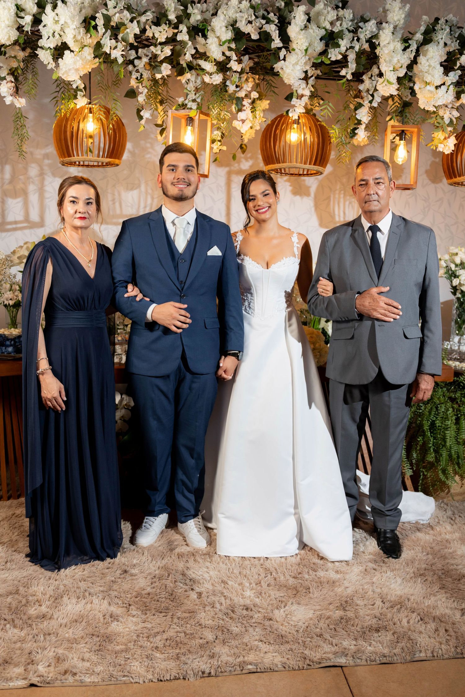 Wedding portrait of bride in white gown and groom in navy suit posing with family under floral decorations.