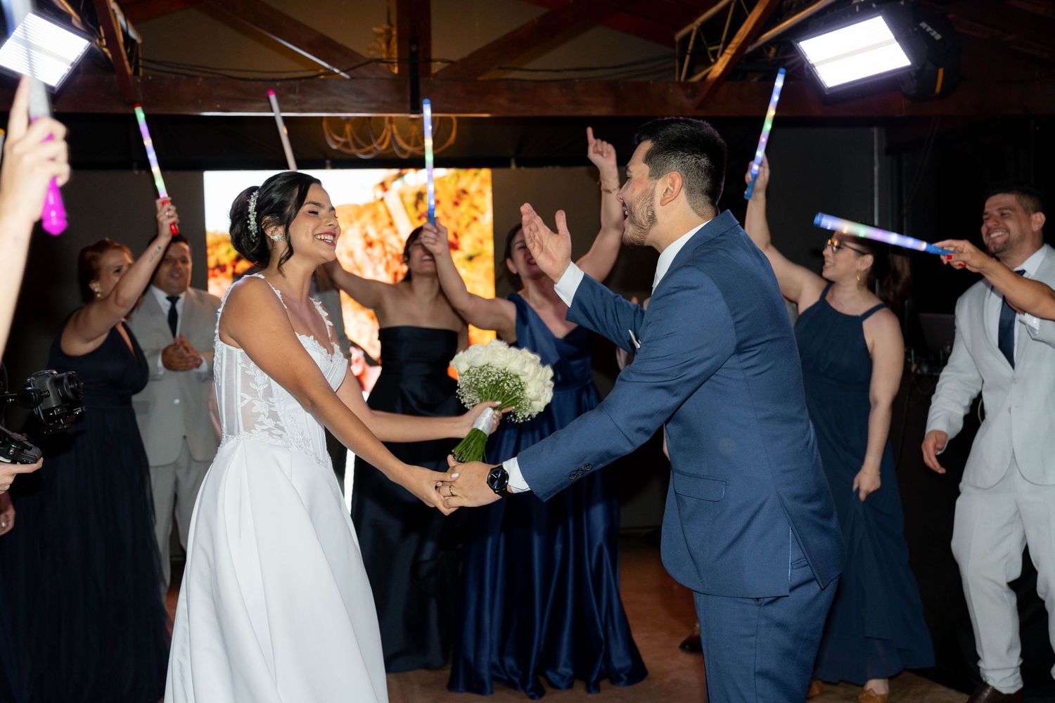 Bride and groom dance joyfully at wedding reception while guests wave glowing sticks in elegant venue.