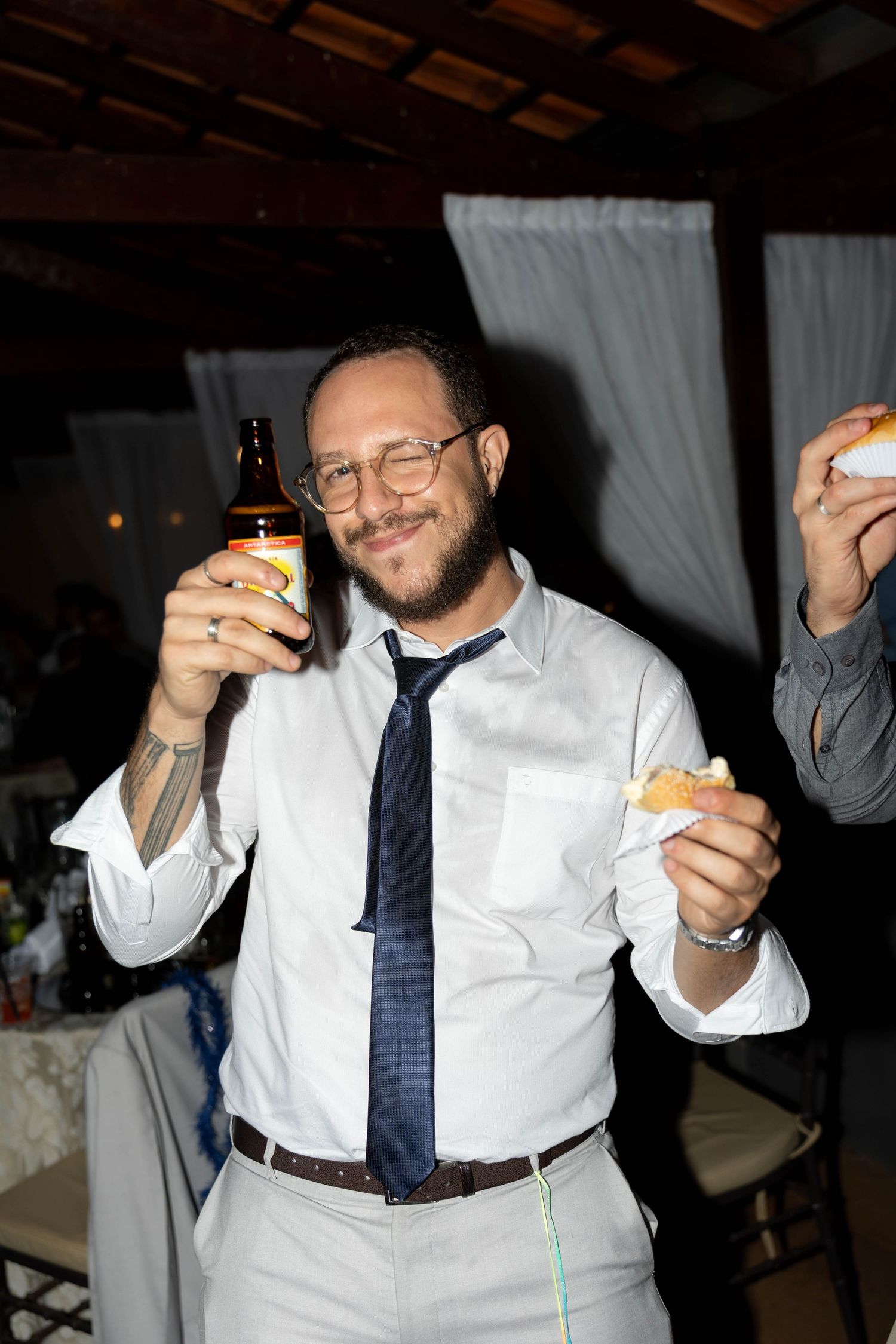 Smiling man in white shirt and tie holding a beer bottle and food at a party event.