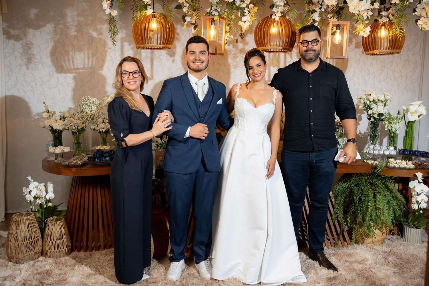 Bride in white gown and groom in navy suit pose with two guests at elegant wedding reception with floral decor.