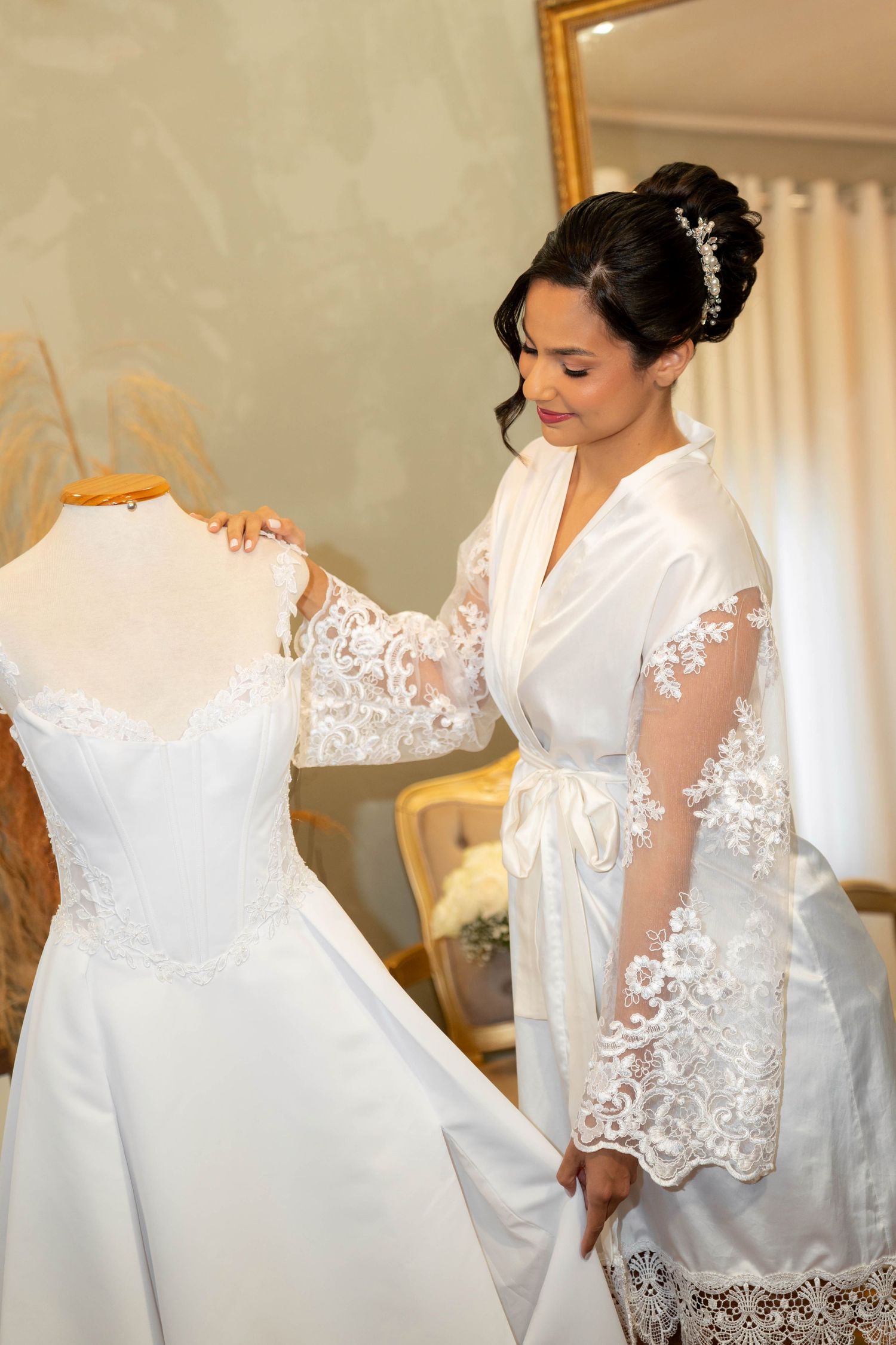 Bride in lace robe admiring her white wedding gown on a dress form in an elegant bridal suite.