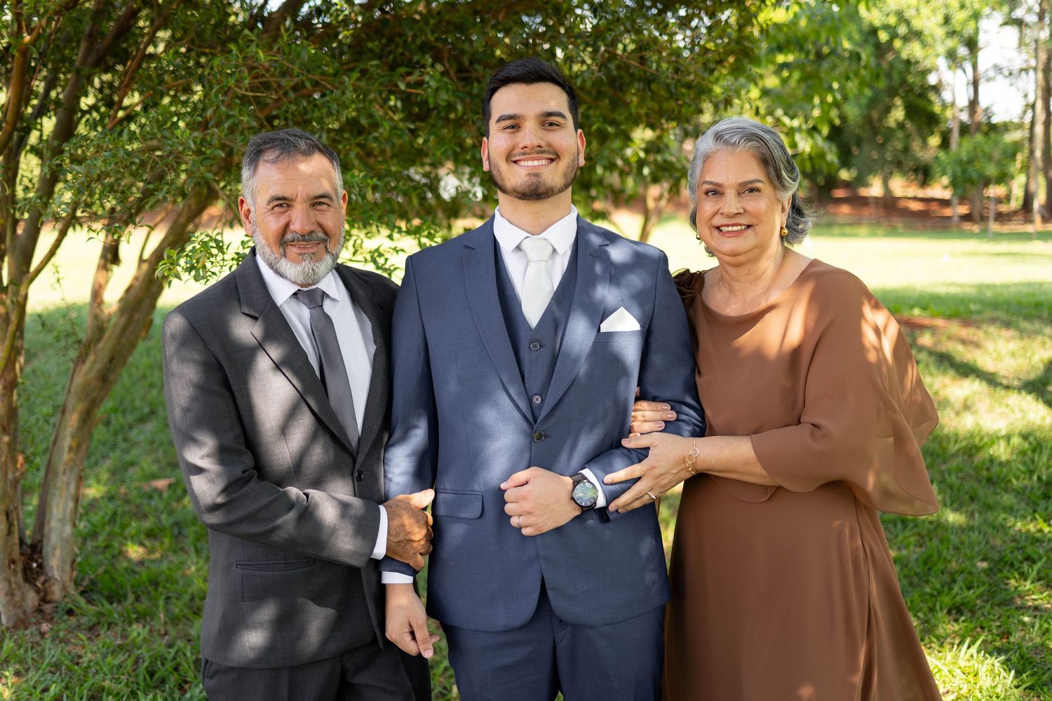 Family of three posing outdoors in formal attire, man in blue suit flanked by older couple, green trees in background.