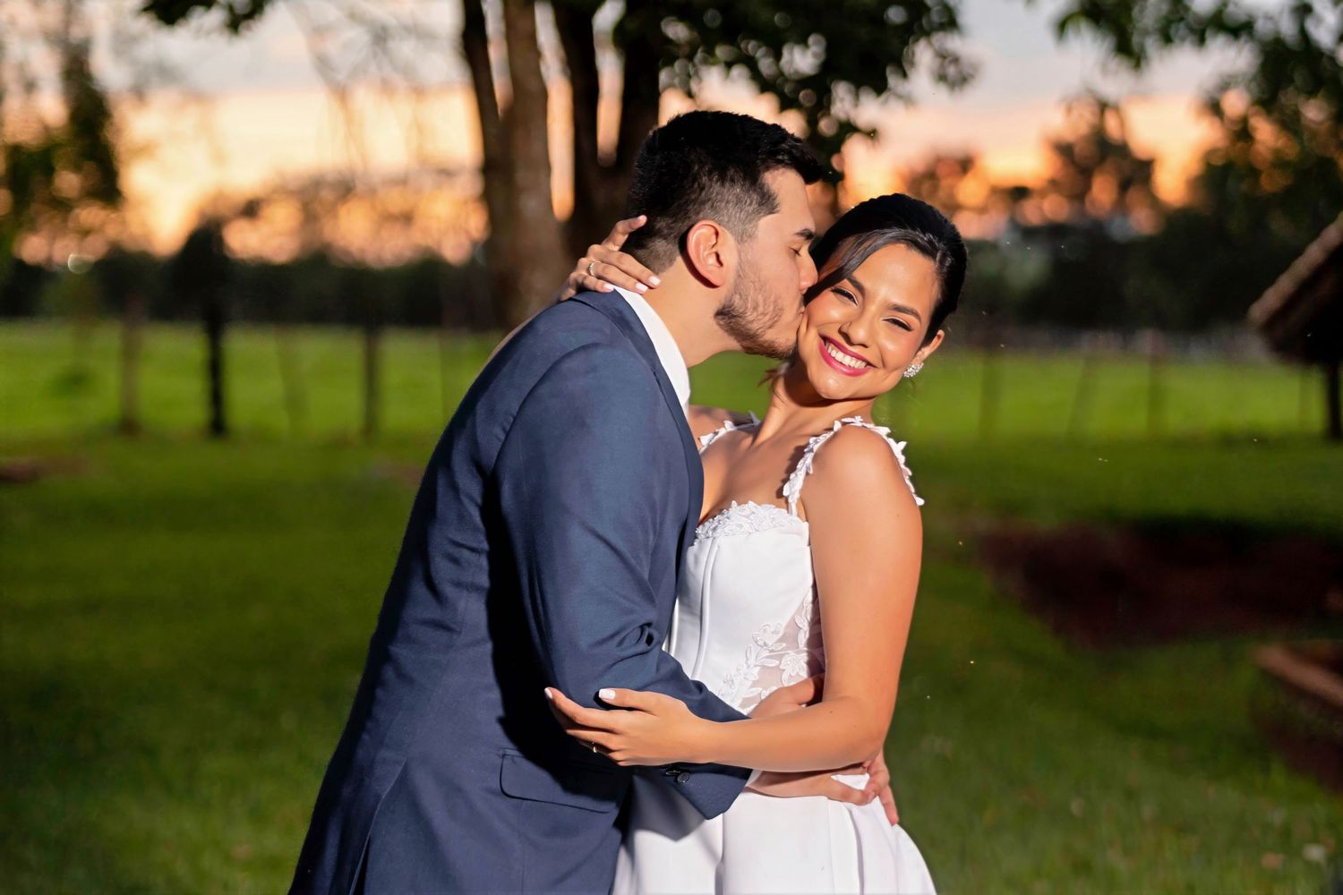 A groom in a blue suit kisses his smiling bride in a white dress outdoors at sunset.