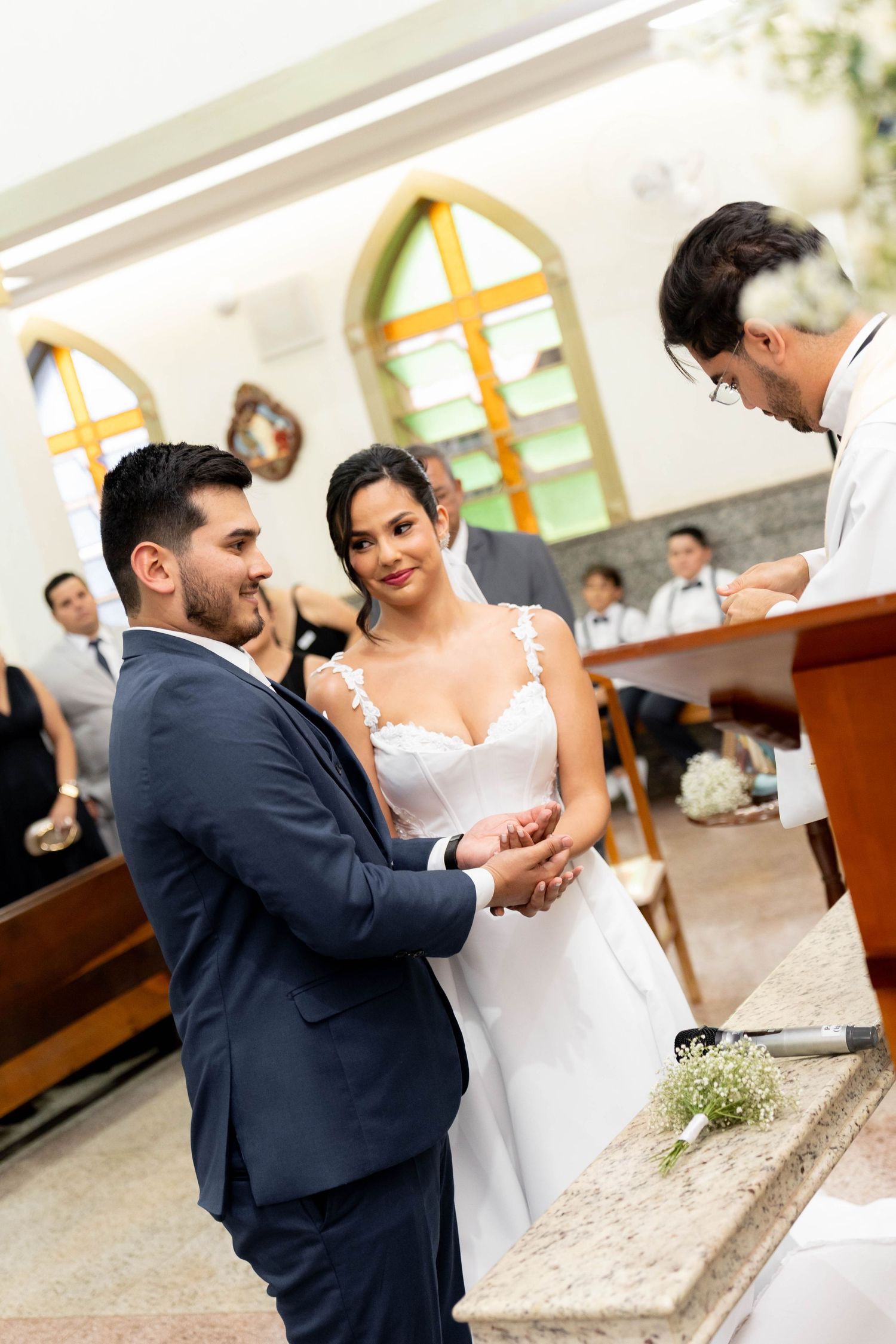 Bride and groom exchange rings at church altar during wedding ceremony, officiant presiding, guests seated in pews behind.