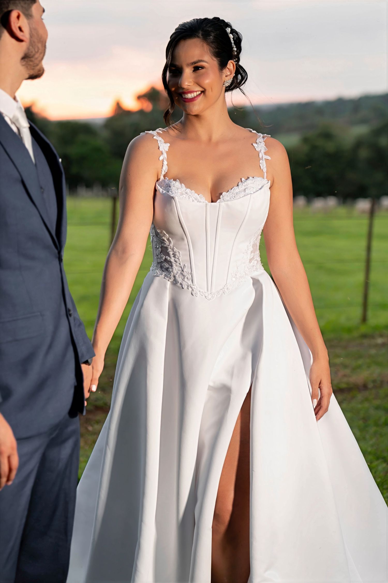 Bride in elegant white corset wedding gown with slit skirt holding hands with groom in gray suit outdoors.