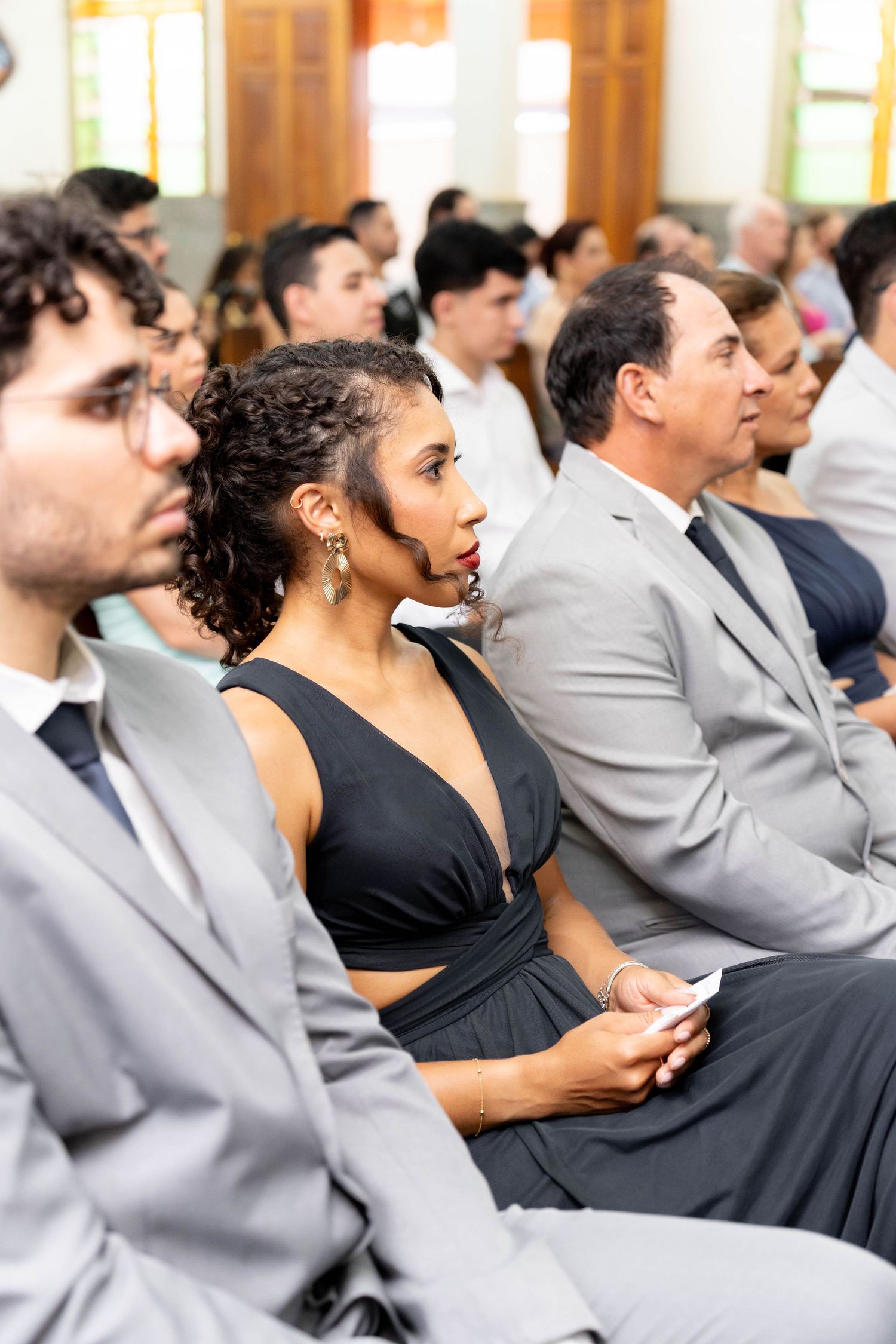 Elegantly dressed guests seated at a formal event, woman in black dress holding phone among men in suits.