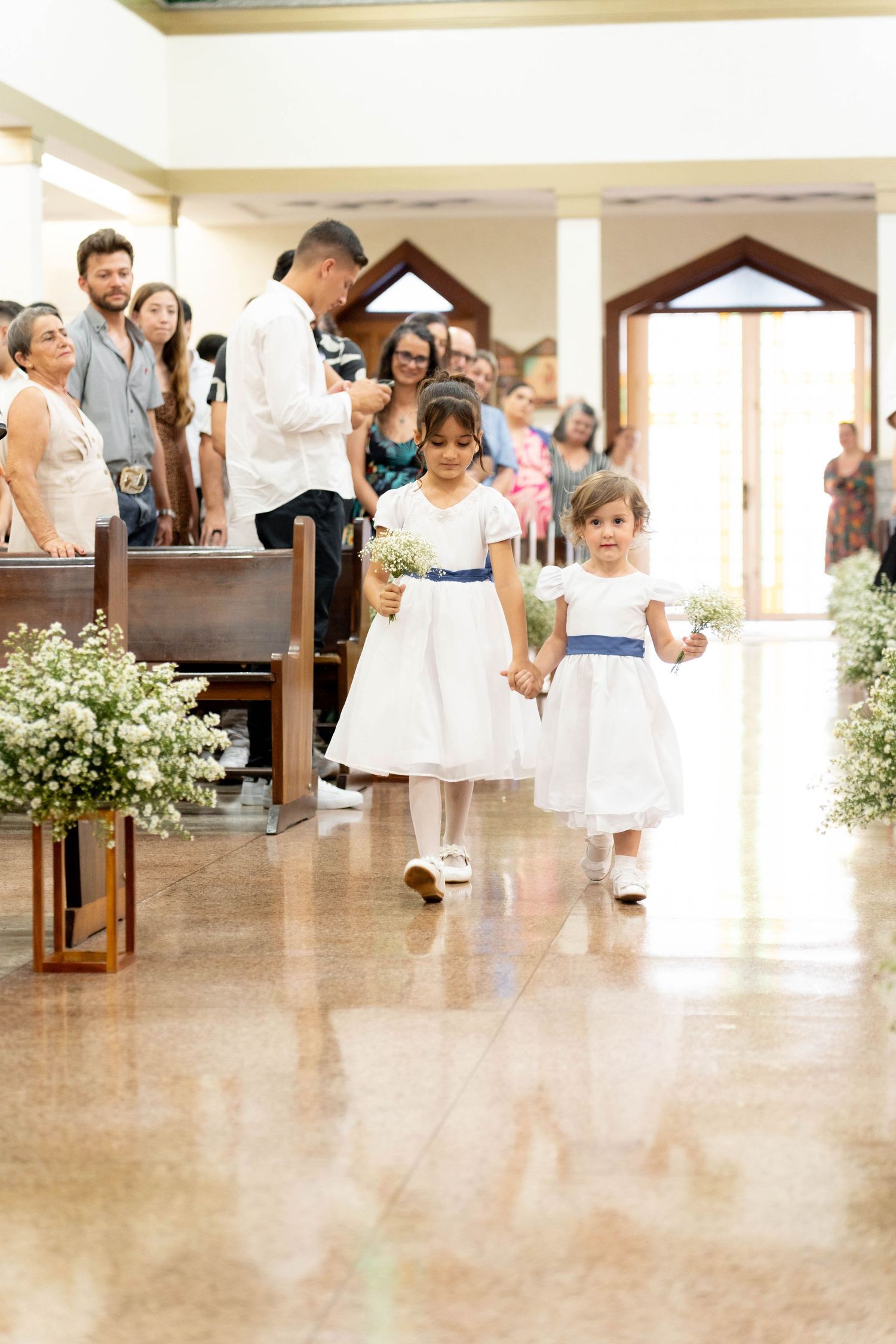 Two young flower girls in white dresses with blue sashes walk down a church aisle at a wedding ceremony.