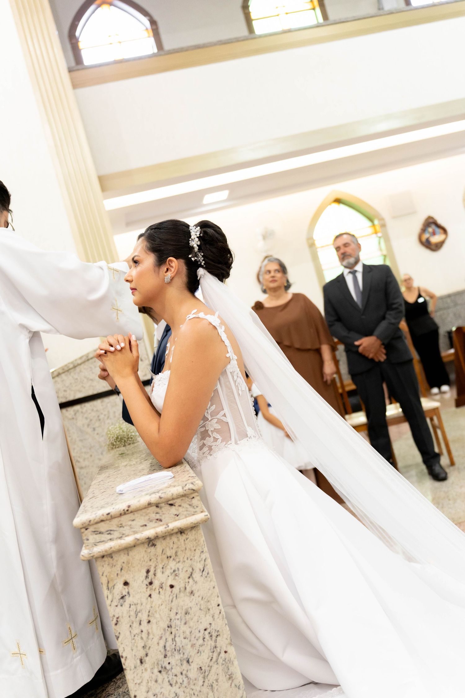Bride in white gown and veil kneeling at church altar during wedding ceremony, guests watching in background.