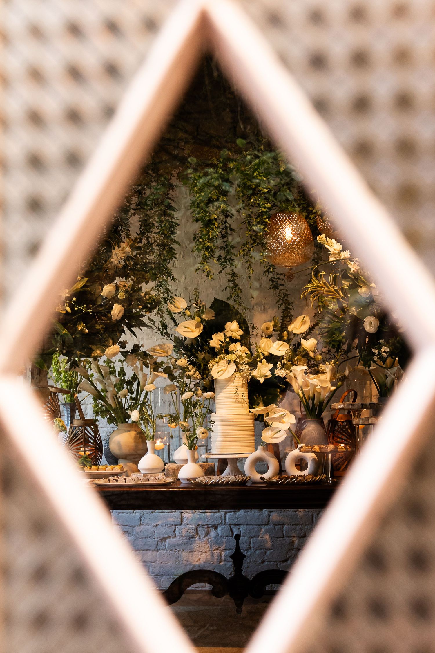Elegant wedding cake display framed by diamond-shaped window, surrounded by white flowers and lush greenery.