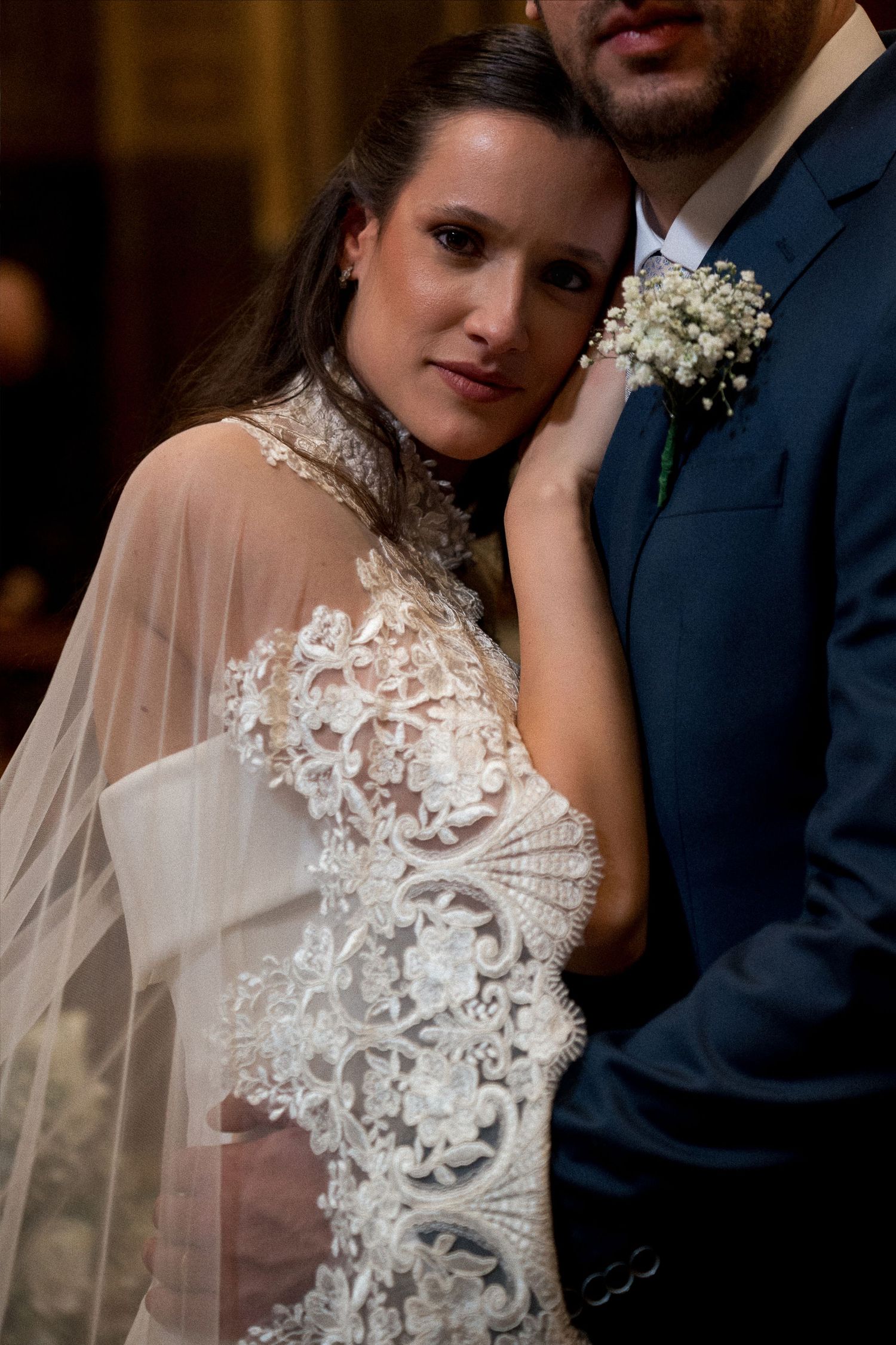 Bride in lace wedding gown with veil leans against groom in navy suit with floral boutonniere.