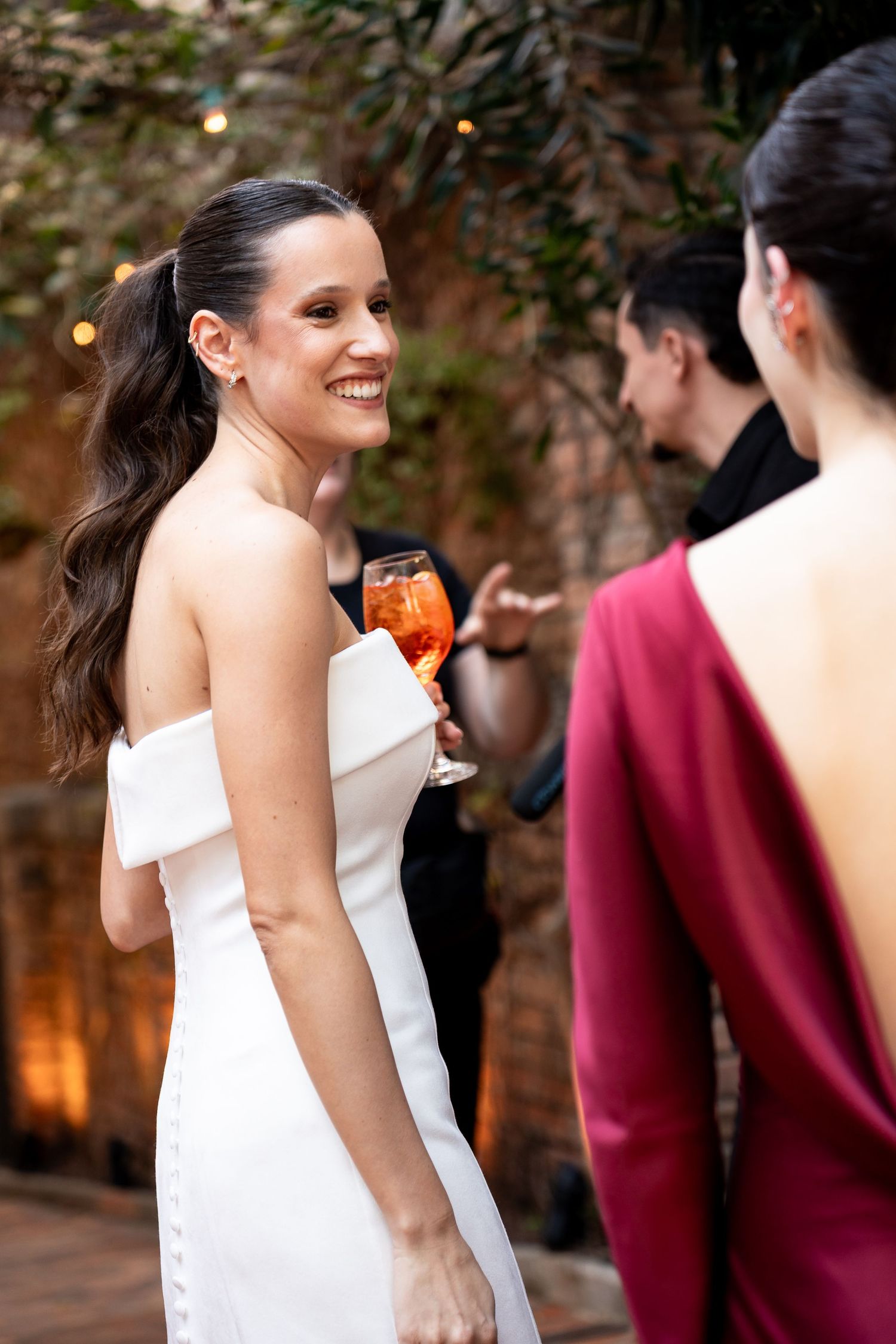 Smiling bride in white off-shoulder gown holds aperol spritz at elegant outdoor wedding reception.
