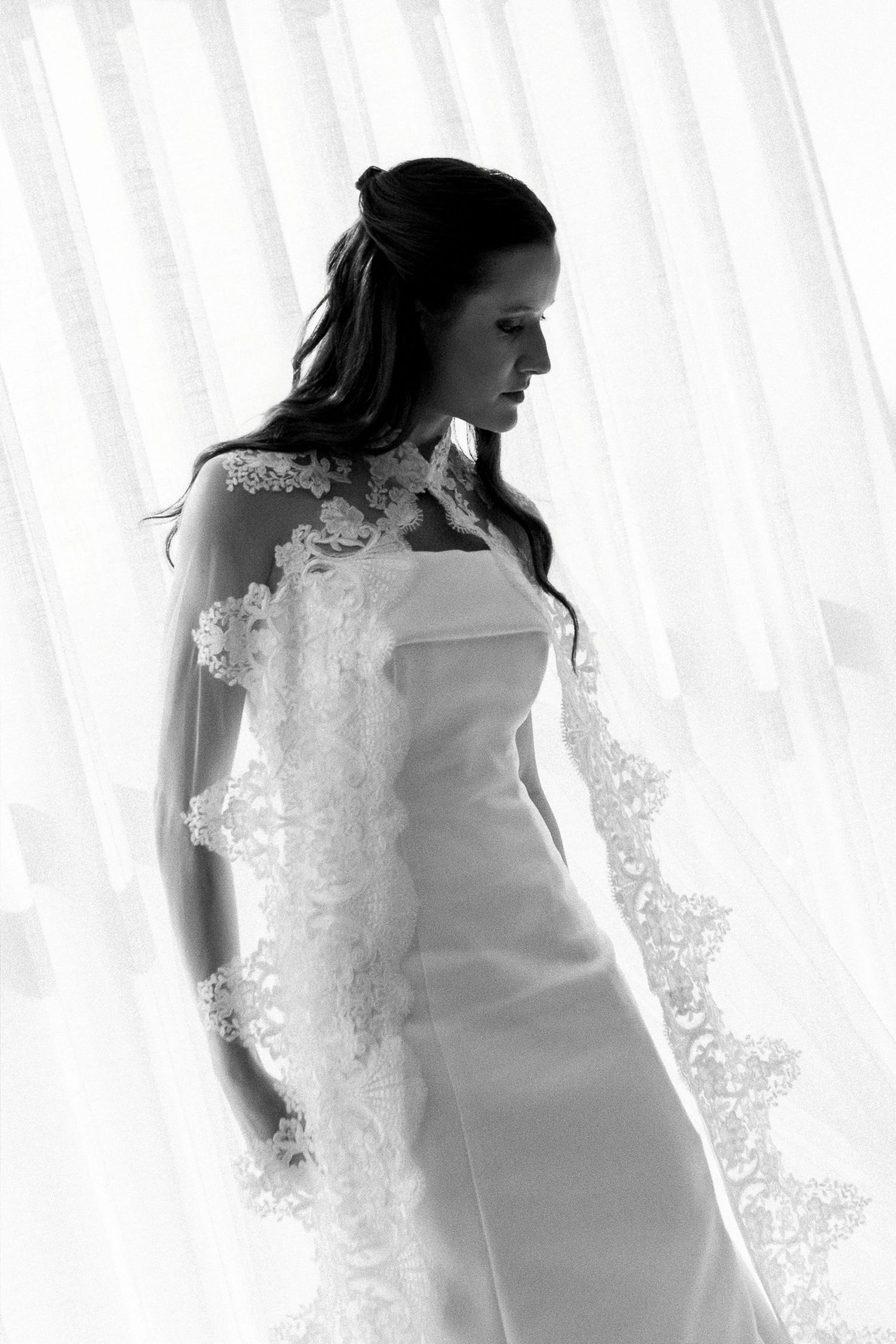 Elegant bride in a white strapless gown with lace veil, posed in black and white near a bright window.